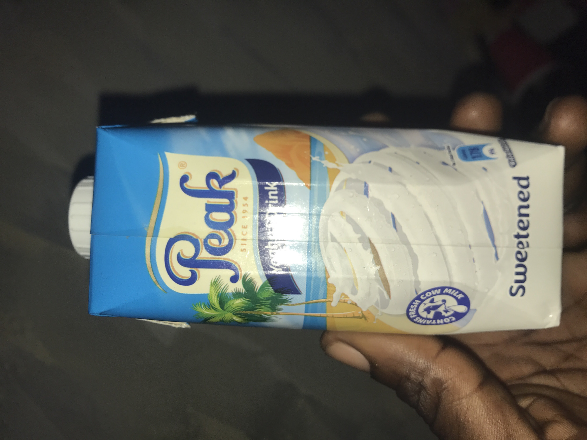 A close-up, horizontal shot shows a hand with dark skin tone holding a rectangular carton of "Peak" brand milk or drink. The carton is predominantly blue on the top and side, transitioning to a light yellow/cream color on the front, with a white side. The "Peak" logo, stylized in blue and gold, is prominent on the blue section, with "SINCE 1954" printed below it. Further to the right, a graphic depicts a beach scene with green palm trees against a blue sky and what appears to be water and sand. A large splash of white liquid, presumably milk, is visible on the white front panel.
Text visible on the carton includes "Peak", "SINCE 1954", and "Drink". On the white side, the word "Sweetened" is printed vertically. A circular blue logo featuring a cow's head and the words "FRESH COW MILK" and "CONTAINS" is also visible. Near the top right of the carton, a small section shows "175ml" and some illegible text. A white screw cap is visible on the left side of the carton. The background is dark and out of focus, suggesting a dimly lit indoor environment. The image is taken in what appears to be evening or night. The overall scene indicates the product is being held, possibly for consumption or display, in Rukuba Road, Nigeria.