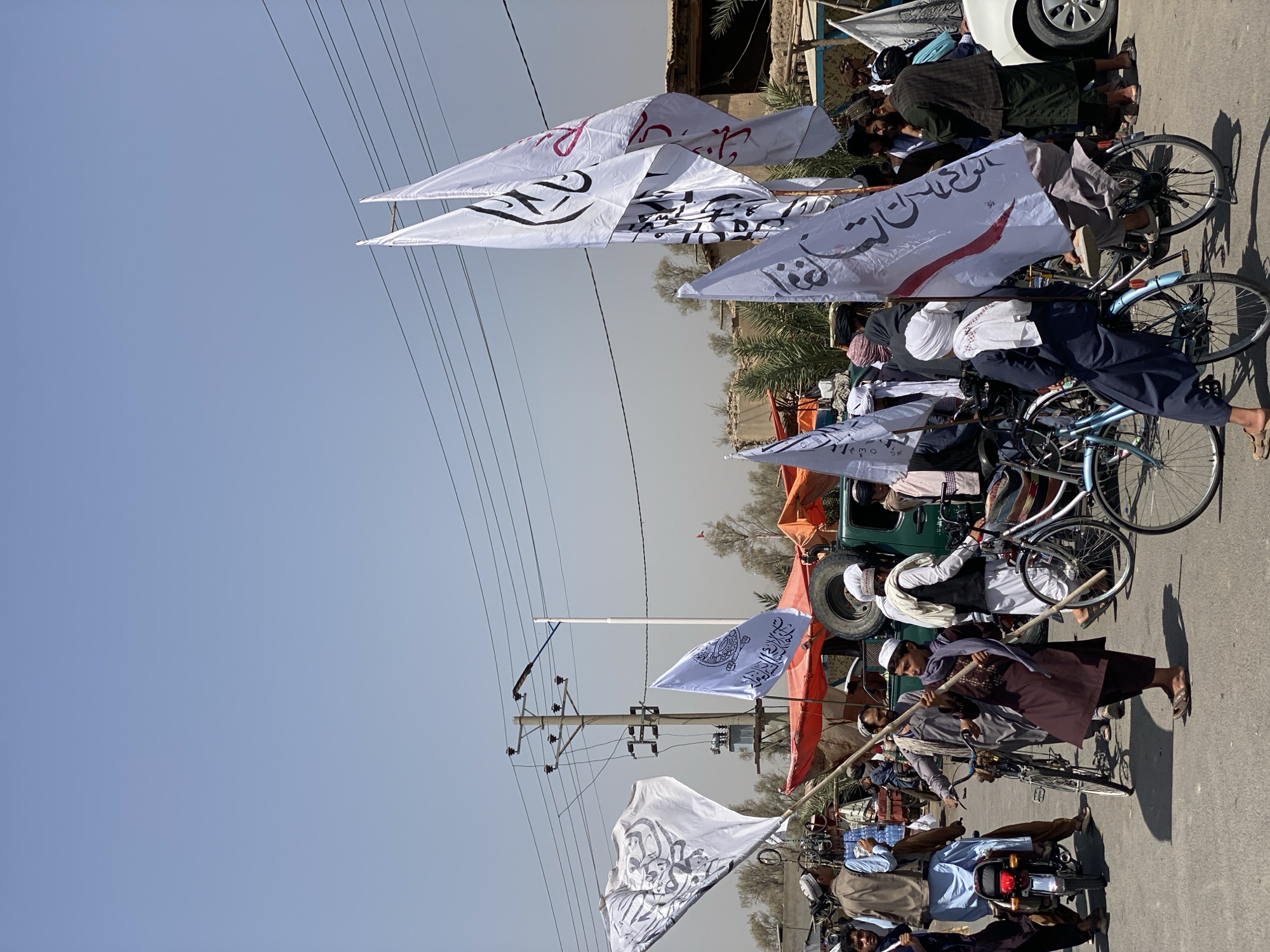 The image depicts a scene in the city of Kandahar, Afghanistan, under a clear, hazy sky. A group of men, many wearing traditional clothing and headwear, are gathered on a paved street. Several white flags with Arabic script are prominently displayed, some attached to poles and others held by individuals. The flags appear to be associated with a particular group or movement.
In the background, a green utility vehicle is parked, along with tents or awnings made of orange and red fabric, suggesting a market or gathering area. Power lines and a utility pole are visible overhead, indicating an urban setting. The presence of motorcycles and bicycles suggests modes of transportation used by the people in the scene. The overall atmosphere appears to be one of a public gathering or event.