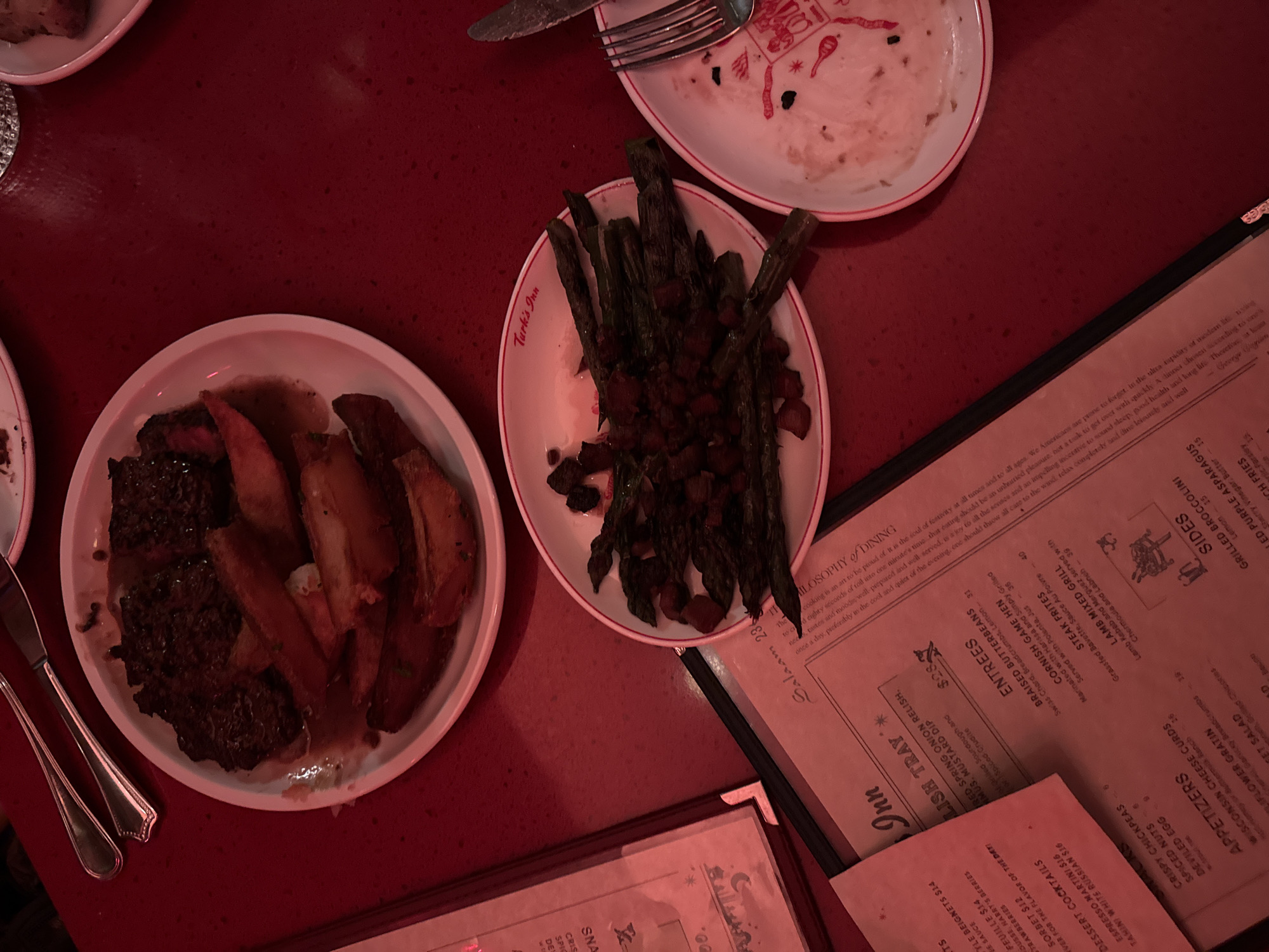 The image depicts a table setting with plates of food and menus at Turk's Inn, a restaurant in New York City, United States of America. From a high angle view, the food includes asparagus with what appears to be bacon bits, steak with potato wedges, and a partially consumed dish. Silverware is also visible, along with menus showing food and drink options. The table has a dark red color, and the lighting appears to be dim.