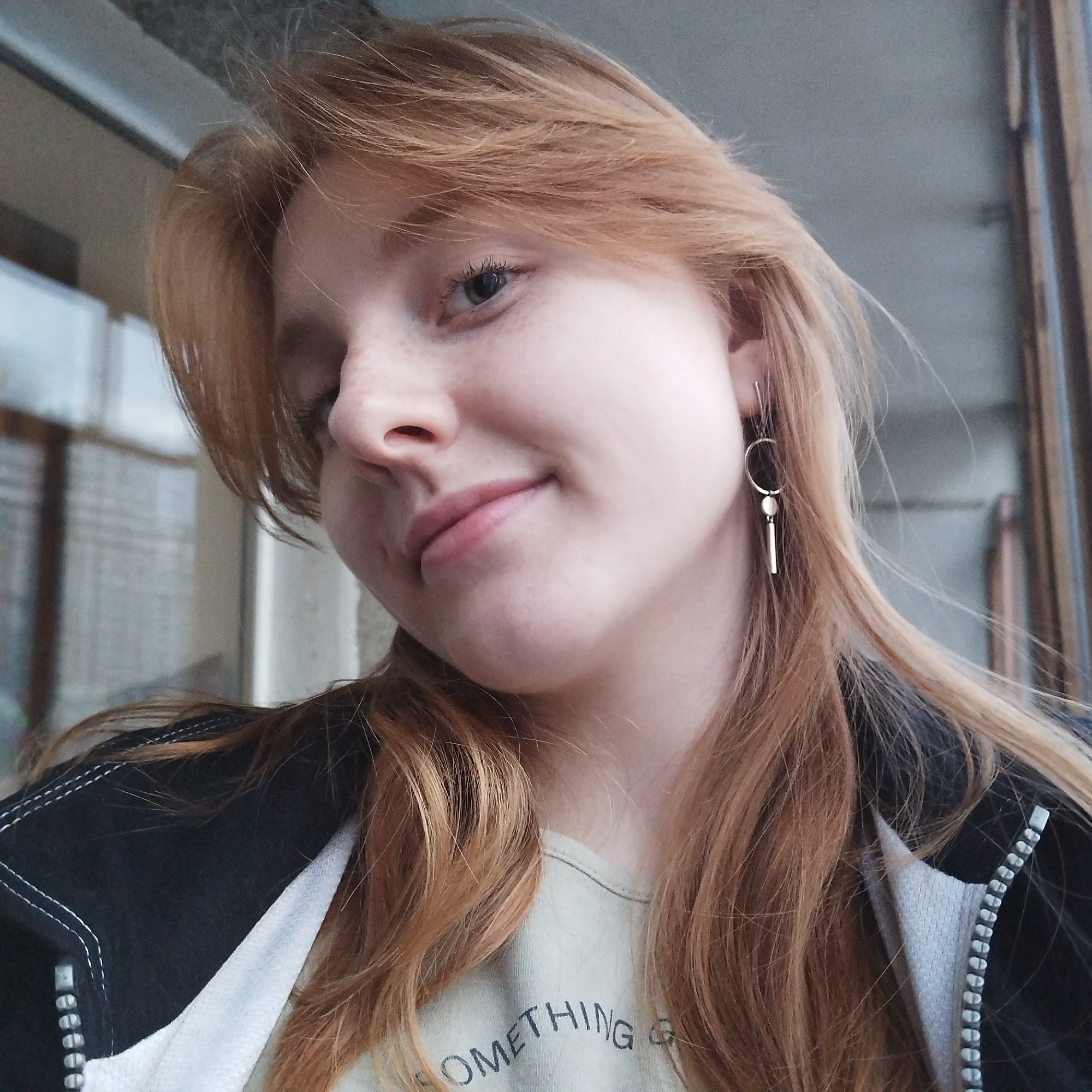 A young woman with red hair is posing for a selfie. She is wearing a black jacket with white trim and a light-colored t-shirt with a partial word visible, beginning with "SOMETHING". She has freckles and is wearing a modern style earring. The background appears to be an outdoor balcony area with a window. The location is unknown and cannot be determined to be Yekaterinburg, Russia.