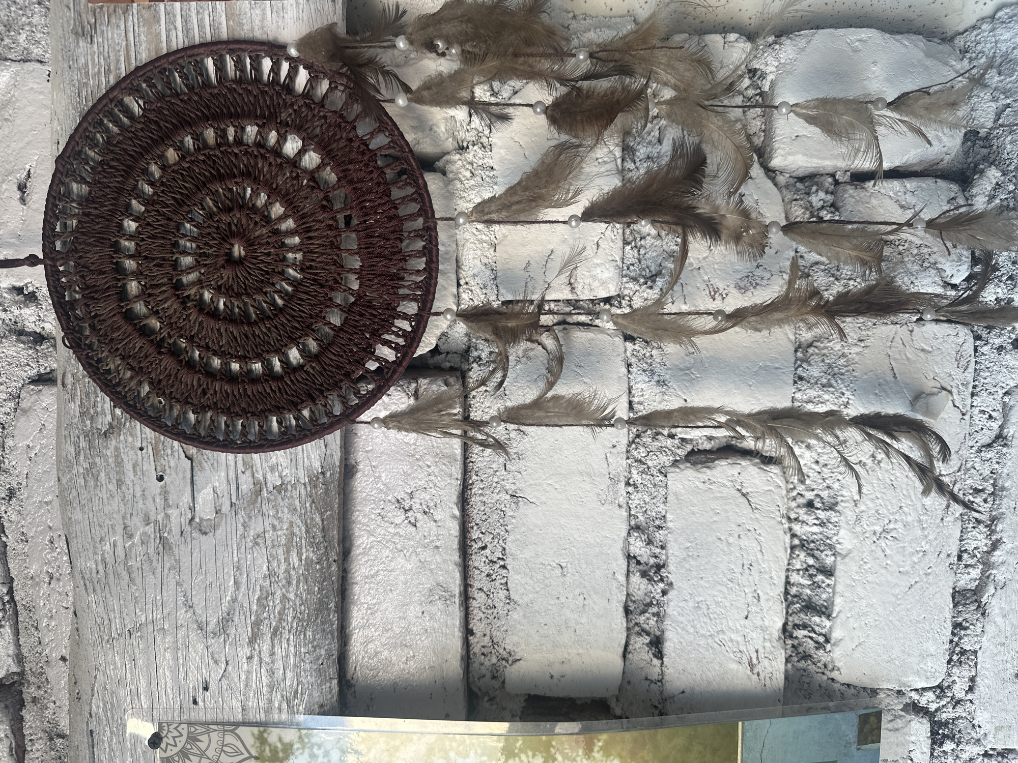 A dreamcatcher hangs against a white brick wall and a weathered wooden beam in Bhagsu, India. The dreamcatcher is circular with a brown, woven pattern and numerous brown feathers hanging from it. Several small white beads are interspersed among the feathers. To the left of the dreamcatcher, a portion of a vertical, framed picture or print is visible, showing a pattern of circles and lines on its left side. The brick wall appears rough and textured, painted white. The wooden beam is horizontal, with a distressed, greyish-white finish showing visible grain and nail holes. The lighting suggests an outdoor or well-lit indoor environment. There are no people or animals visible in the scene.