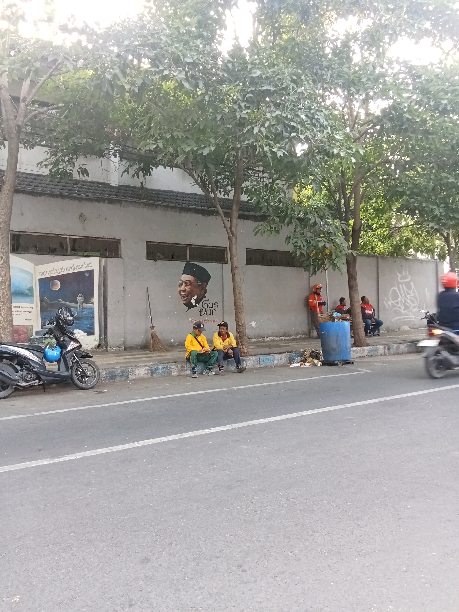 The image shows a street scene in Kaliwungu, Indonesia, with a building on the left adorned with murals. One mural depicts an open book with an astronaut on a lunar surface, and another features a portrait of a man wearing a peci hat and glasses, with the name "Gus Dur" written below him.
In front of the building, a black motorcycle is parked. Two men in yellow shirts and green pants are sitting on the curb, facing the street. A third man, also in yellow, is sitting to their right. Further down the sidewalk, a man in an orange jacket stands next to a blue trash bin filled with debris. Two other men are seated near the trash bin. A motorcycle with a rider wearing a red helmet is moving on the street from right to left. The environment includes trees and a gray wall with graffiti. The road is paved with asphalt and has white lane markings. The lighting suggests it is daytime, possibly late afternoon, with sunlight filtering through the trees.