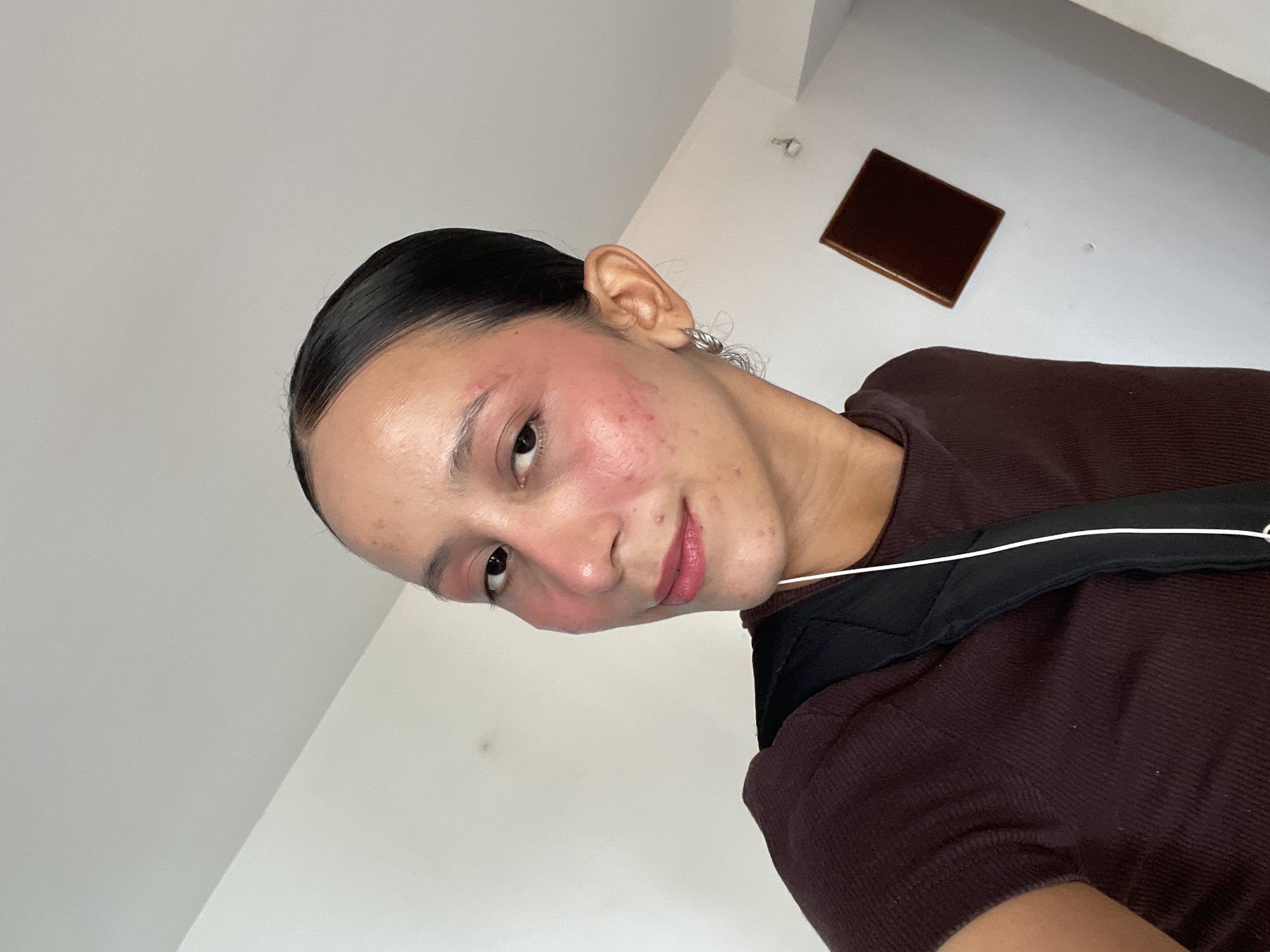 A young woman with dark, pulled-back hair and a brown shirt is looking towards the camera. She is wearing makeup, including blush on her cheeks and pink lipstick. She has earrings in her ears and appears to be wearing headphones. A black backpack strap is visible across her shoulder. The background is a plain, white wall with a brown rectangular object mounted on it, and a shadow is cast on the wall. The lighting suggests an indoor setting, possibly during the day. There is no information about the city of Porlamar, Venezuela, or any specific location within it.