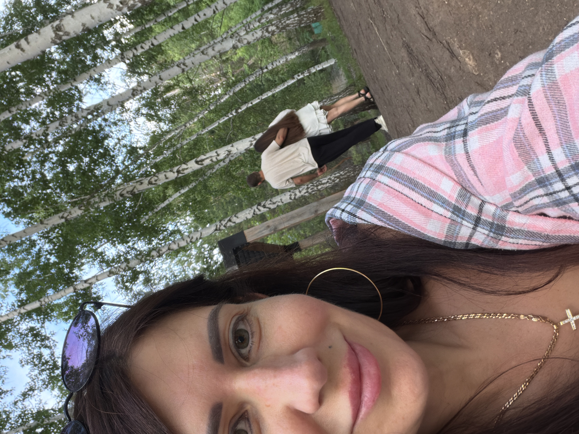 In Volzhskiy, Russia, a woman with long dark hair and sunglasses perched on her head takes a selfie. She is wearing a pink and grey plaid shirt, a gold chain with a cross pendant, and large hoop earrings. In the background, a couple walks arm-in-arm through a birch forest. The man is wearing a light-colored t-shirt and dark pants, and the woman is in a white dress. The ground is covered in dirt and fallen leaves, with dappled sunlight filtering through the trees.