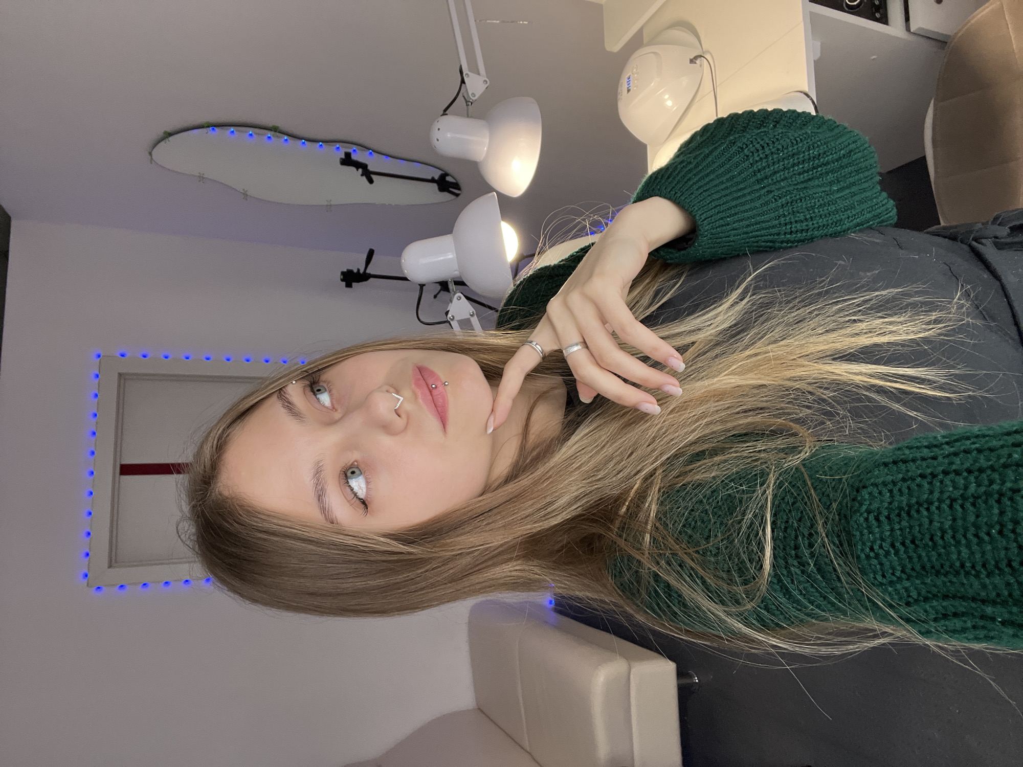 A young woman is indoors. She has light skin, light brown hair, and green eyes. She is wearing a dark shirt and a dark green knitted hat or scarf. She has piercings on her nose and lip, and rings on her fingers. Behind her is a white wall with a rectangular outline of blue lights, a white ceiling, white lamps, and a white salon table. The location is Фоки, Russia.