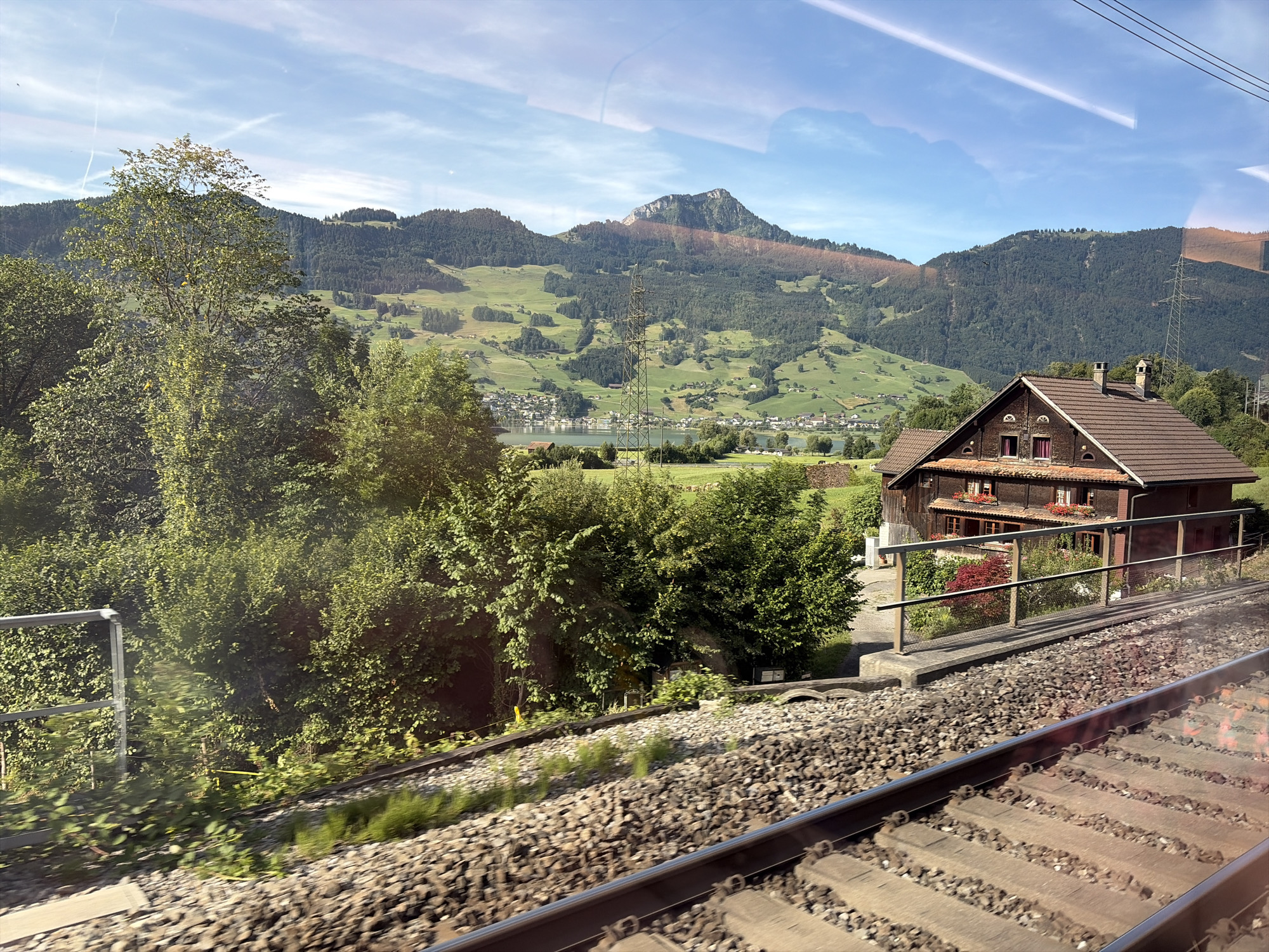 This is a photograph taken from a moving train, offering a view of the Swiss countryside near Steinen, Switzerland. The foreground is dominated by railroad tracks and ballast, with a metal railing and lush green bushes along the side. In the midground, a traditional wooden house with a red-tiled roof and multiple chimneys is situated on a hillside. Red flowers adorn the windowsills of the house. Beyond the house, a body of water, likely a lake, is visible, with a small town or village on its shores. Further in the background, rolling green hills, covered in patches of dark green forest and lighter green fields, rise up to meet a majestic mountain range. The sky is blue with wispy white clouds and faint contrails from an aircraft. The overall impression is a picturesque, rural landscape under a bright, sunny sky.