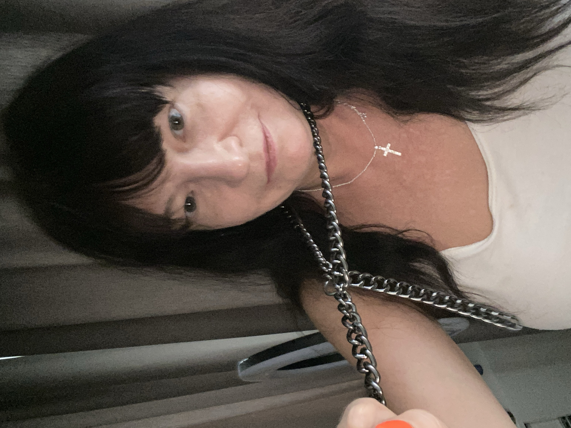The media file is a close-up, self-portrait of a woman. She has long, dark hair with bangs and is wearing a light-colored tank top. A silver necklace with a cross pendant is visible around her neck. She is also wearing a dark, chunky chain, possibly part of a purse strap, which is held in her right hand. Her expression is a gentle smile, and her eyes are looking towards the camera. The background appears to be a dimly lit interior, with horizontal blinds or some textured surface visible. The orientation of the photo suggests it was taken with a smartphone held at an angle. The subject's posture and facial expression convey a relaxed and casual mood. No text is visible in the image.