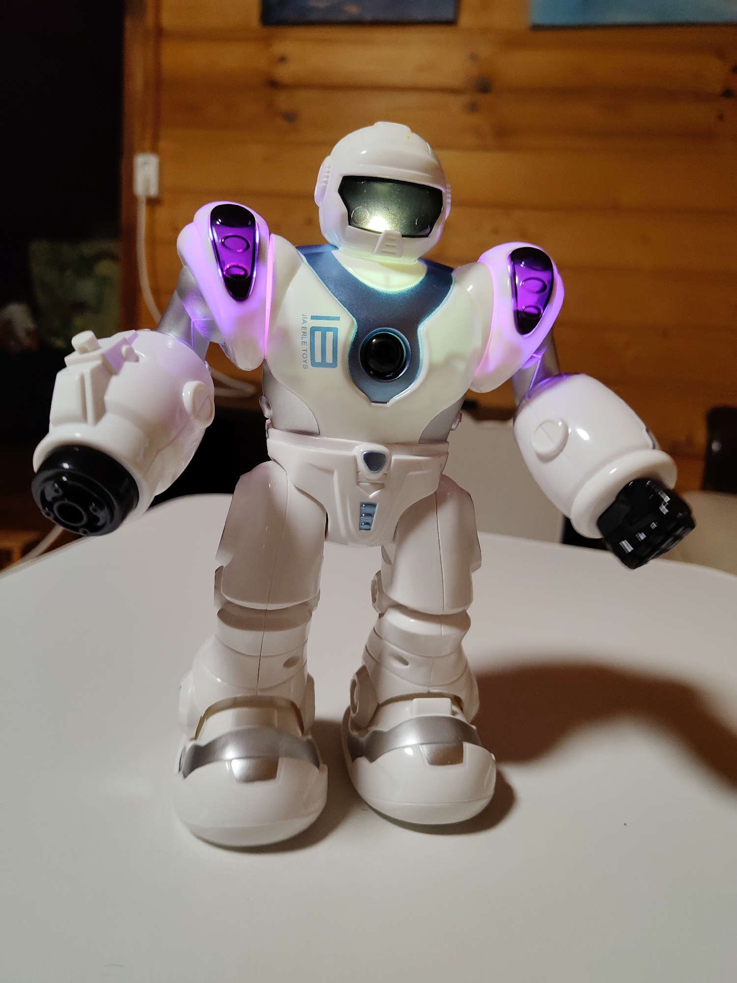 A white and blue toy robot stands on a white table. The robot has purple lights on its shoulders and green lights on its chest. The background is a wooden wall. The robot is approximately 12 inches tall. The scene is located in the city of Фестивальное Снт, Russia.