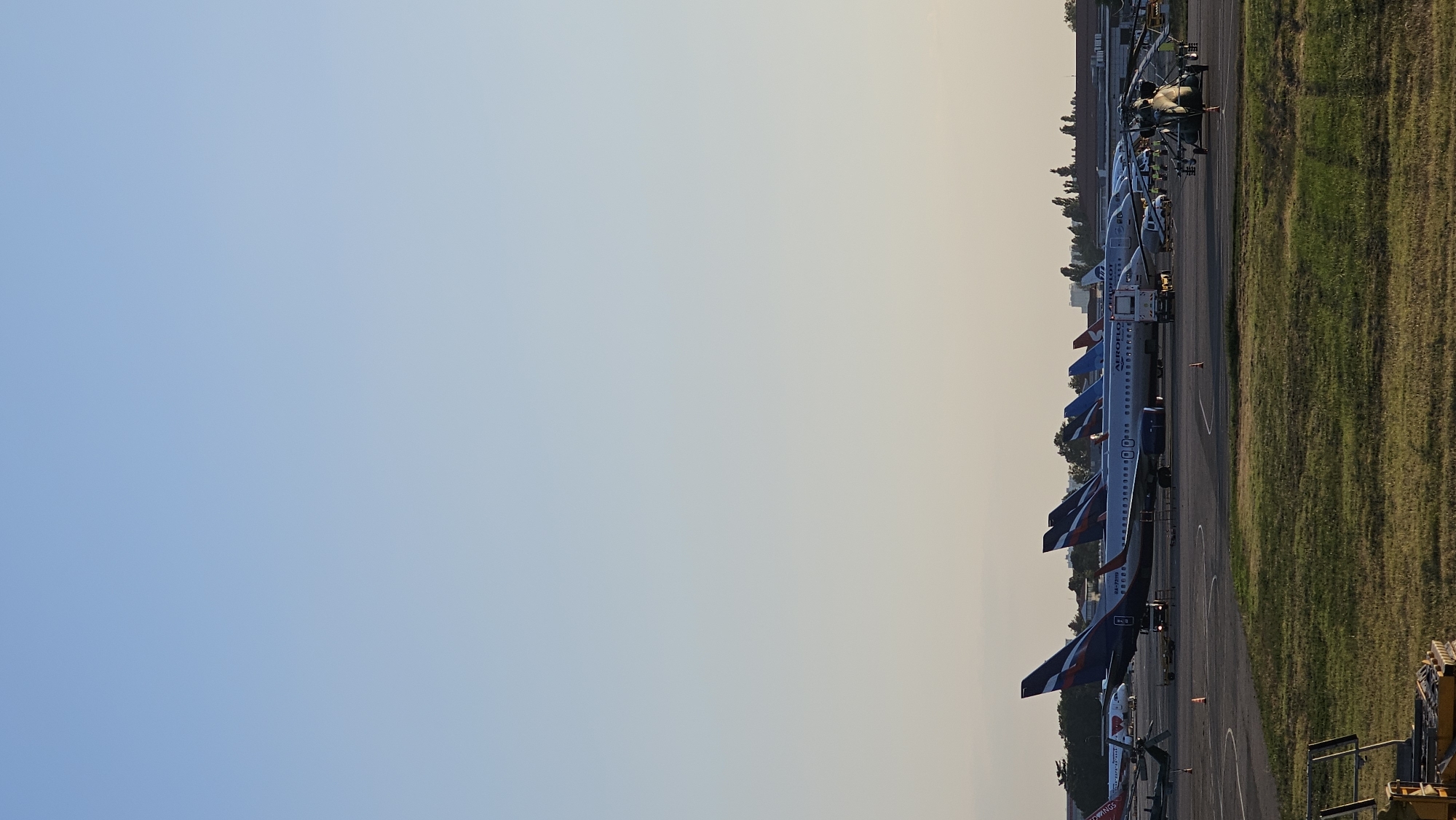 The image depicts an outdoor scene at an airport, likely in Krasnodar, Russia, given the presence of Aeroflot aircraft. Several passenger airplanes are parked on the tarmac, with their tails prominently displaying the Aeroflot livery. Some of these aircraft appear to be lined up. In the foreground, a helicopter is visible, positioned slightly to the right of the frame. The ground is paved in some areas and covered with green grass in others. In the background, there are buildings and trees, suggesting an airport infrastructure. The sky is clear and exhibits a gradient of light blue to pale yellow, indicating either early morning or late afternoon lighting conditions. The overall scene is static, with no overt actions or interactions taking place.