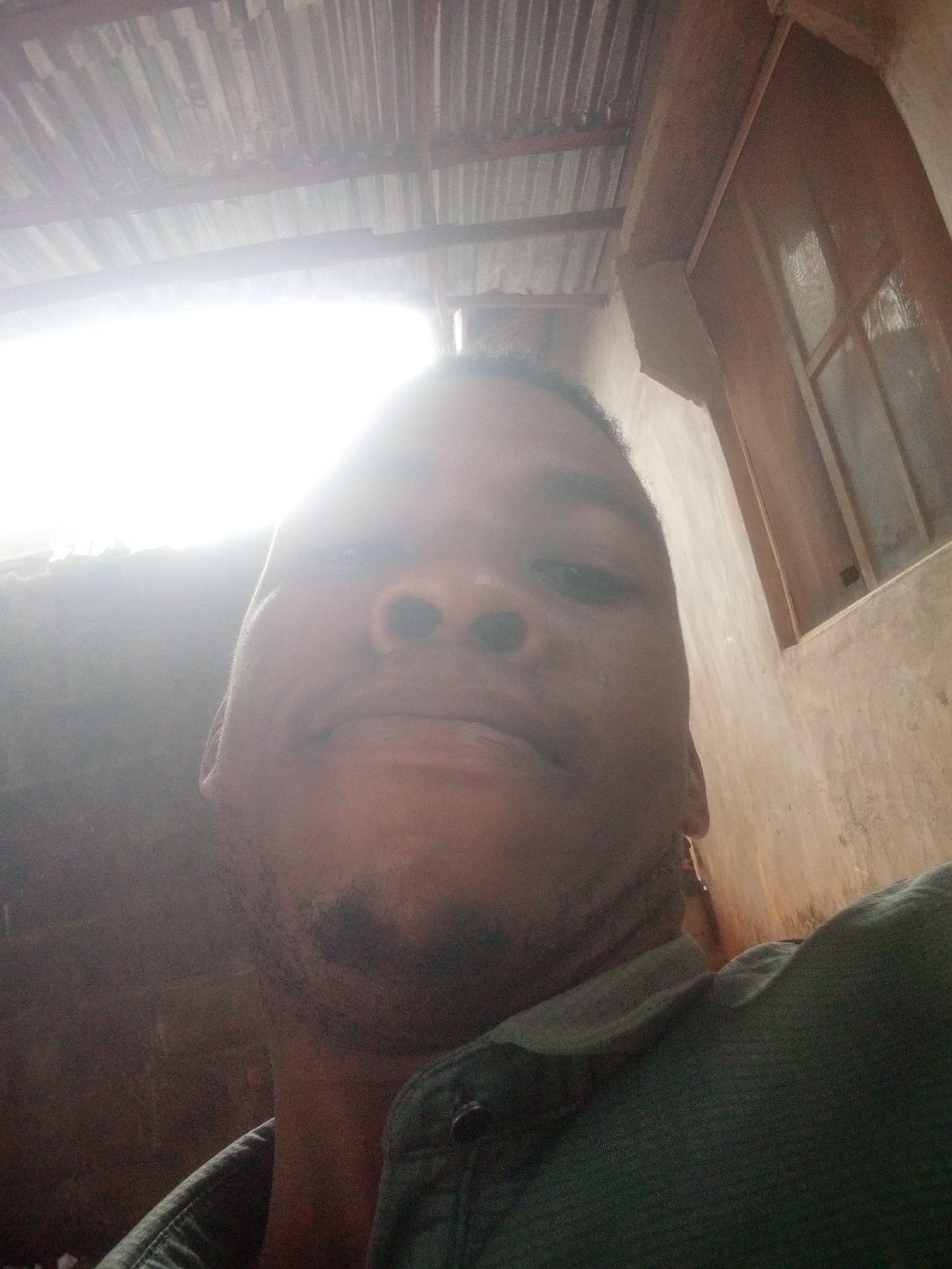 A close-up selfie of a person with dark skin is presented. The person has a mustache and a short beard. They are wearing a green shirt with a button visible near the collar. The background is indistinct, but there is a corrugated metal roof and a window frame, along with a wall. The location is Unknown, Unknown. There is strong light coming from above, causing glare.
