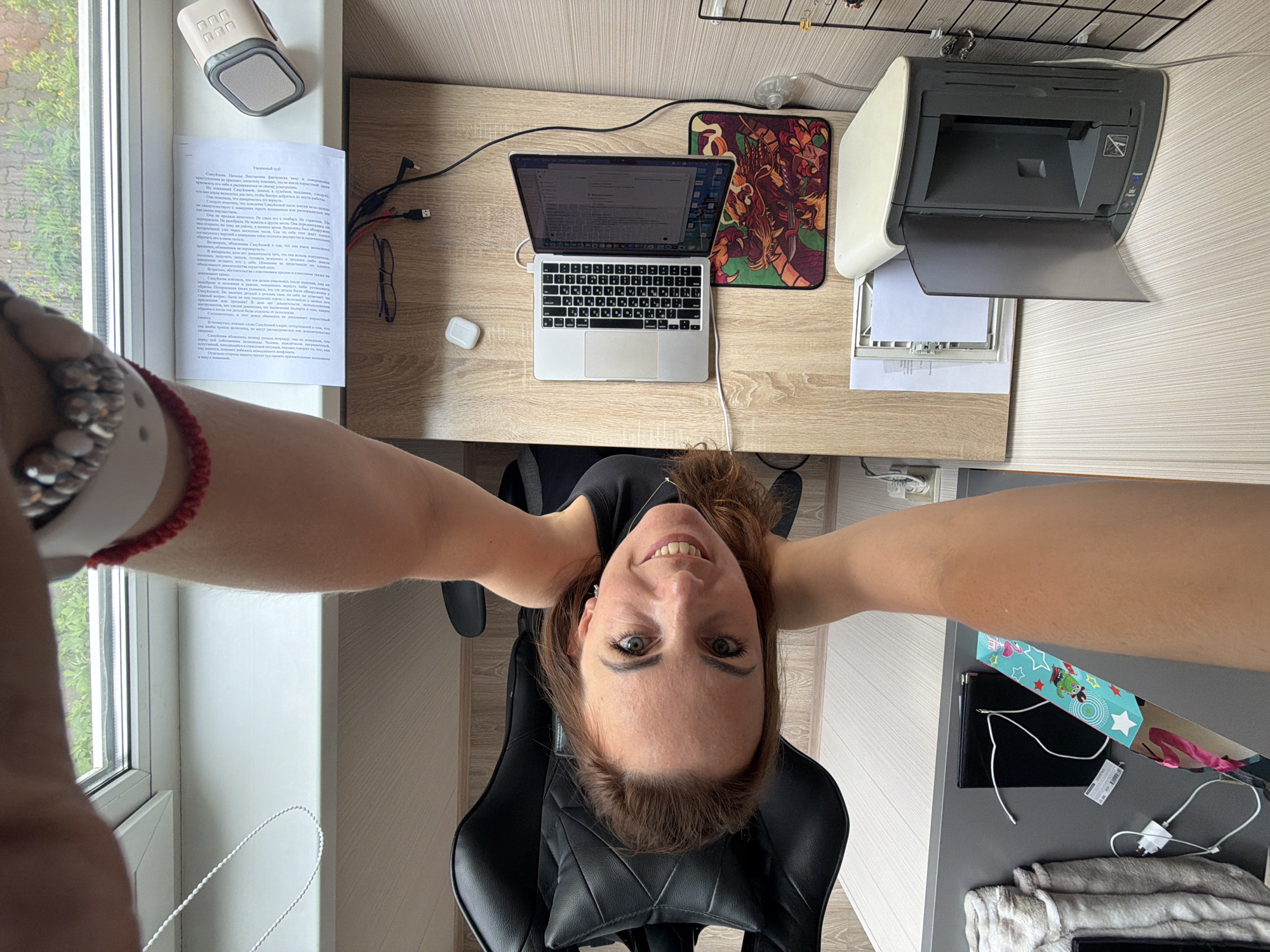 A woman is seated in a black office chair at a light wood desk. She is wearing a black top. Her head is tilted back, and she is looking up at the camera, smiling. Her right arm is bent at the elbow, and her hand is positioned near her face. On the desk, to her right, is a silver laptop, open and displaying text. A colorful dragon-themed mousepad is beneath the laptop. To the left of the laptop is a white wireless earbud case. Behind the laptop, on the desk, are a pair of glasses, and some cables. Further back on the desk, near the window, is a sheet of paper and a small beige speaker. To the right of the desk, on a lower surface, is an open printer. The scene appears to be a home office or workspace, with natural light coming from a window above the desk. The overall impression is one of a casual, perhaps slightly playful, interaction with the camera.