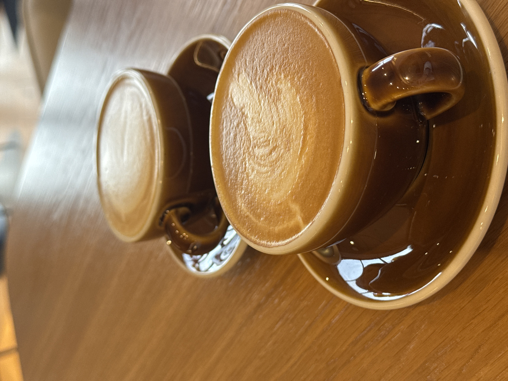 Two brown ceramic cups of coffee, each with latte art, sit on matching saucers on a wooden table. The cup in the foreground is in sharp focus, showing the creamy texture of the foam and a swirling pattern. The second cup is slightly out of focus in the background. The lighting suggests an indoor setting, possibly a cafe. There are no people visible in the image.