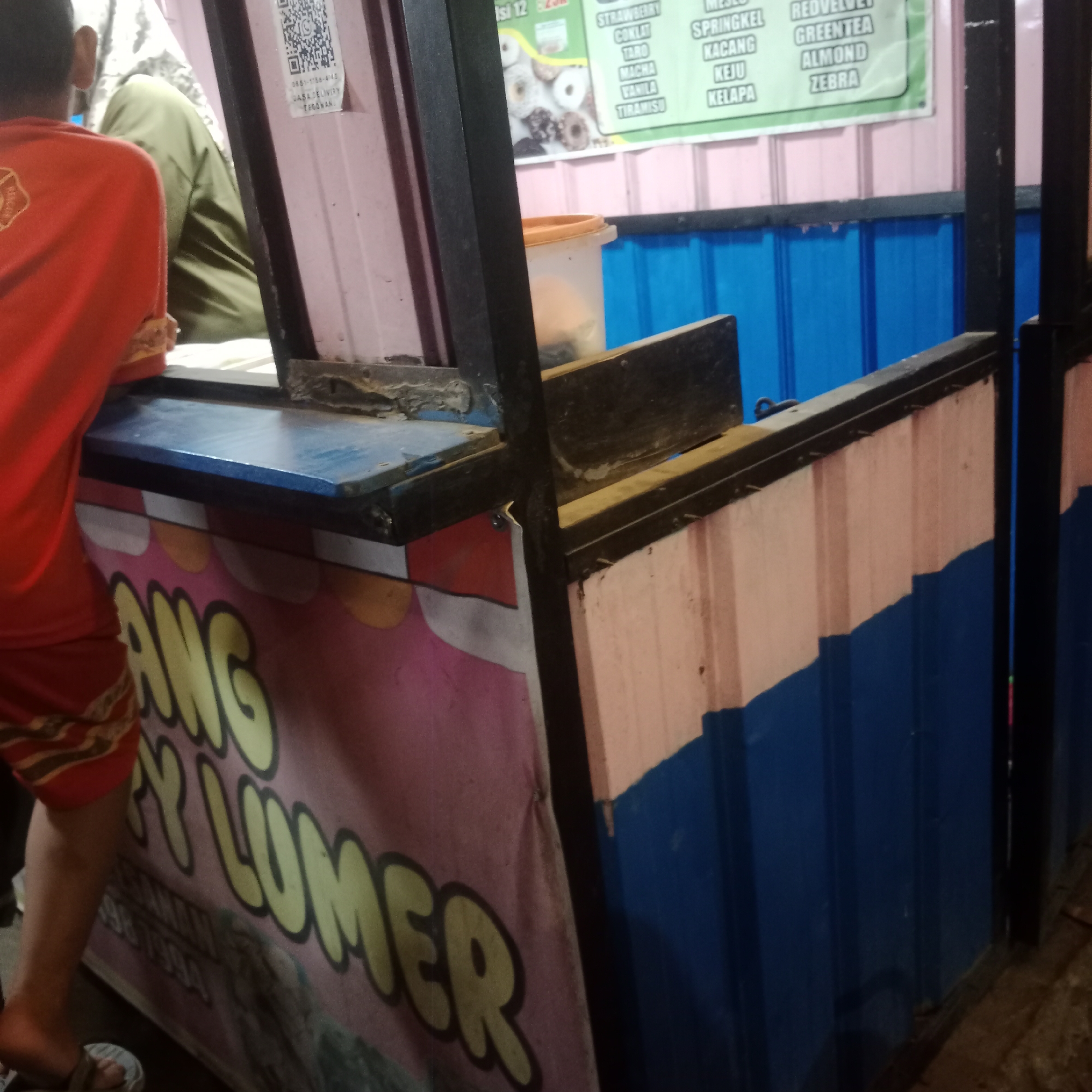 The image depicts a food stall, possibly a dessert stand, in Tegowanu Kulon, Indonesia. A person wearing a red shirt and shorts is partially visible on the left, facing away from the camera and standing near the stall's counter. The stall has a pink and yellow striped front with the text "ANG Y LUMER" written in large yellow letters, likely the name of the business. Above the counter, there is a menu board listing various flavors such as "STRAWBERRY," "COKLAT," "MACHA," "VANILA," "TIRAMISU," "SPRINGKEL," "KACANG," "KEJU," "KELAPA," "REDVELVET," "GREENTEA," "ALMOND," and "ZEBRA." A plastic container with a lid is visible on the counter. The stall structure appears to be made of metal and colored panels, with a blue corrugated metal base visible in the background. The overall scene suggests an outdoor food vendor operating in the evening or at night, as indicated by the lighting.