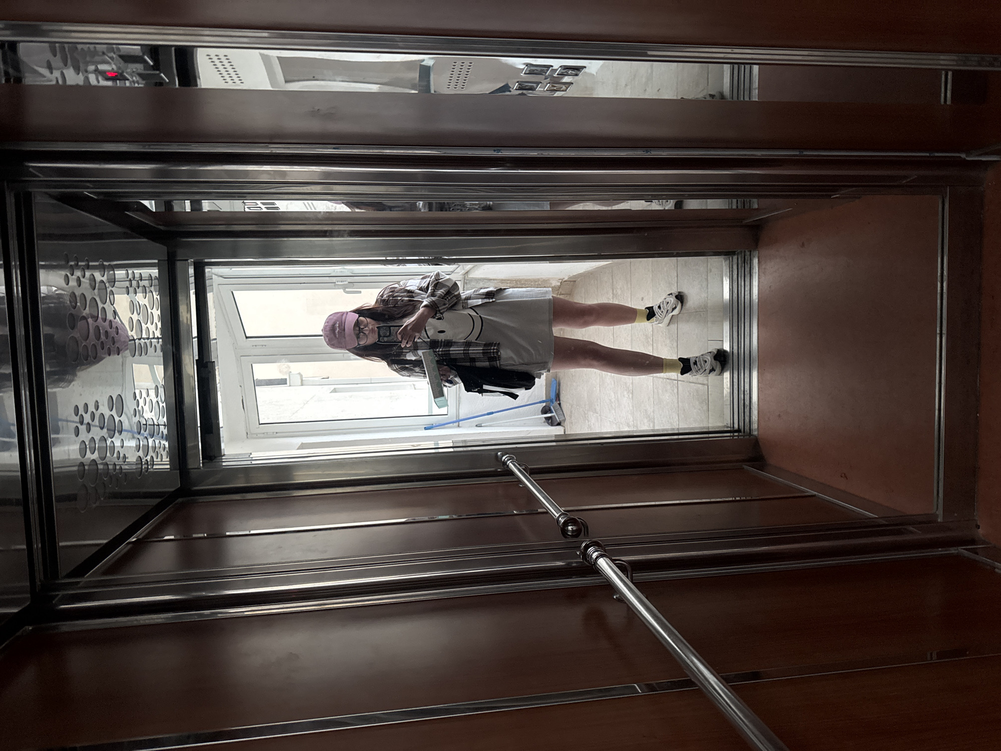A person is seen taking a selfie in a highly reflective environment, likely an elevator or a corridor with mirrored walls. The individual, a young woman with long dark hair and glasses, is wearing a pink baseball cap, a plaid shirt over a light-colored t-shirt with a smiley face graphic, a light-colored skirt or shorts, and white sneakers with yellow socks. She holds a smartphone with both hands, directing it towards her reflection.
The setting is characterized by dark wooden panels interspersed with polished stainless steel accents, including horizontal handrails along one side. The reflective surfaces offer glimpses of the surrounding environment: a bright outdoor scene viewed through a window or glass door, a light-tiled floor, and a blue-handled broom. A small, red digital display, possibly indicating the number "1," is also reflected on an upper panel, alongside decorative circular patterns on another reflective surface. The bright light from the reflected exterior suggests it is daytime.