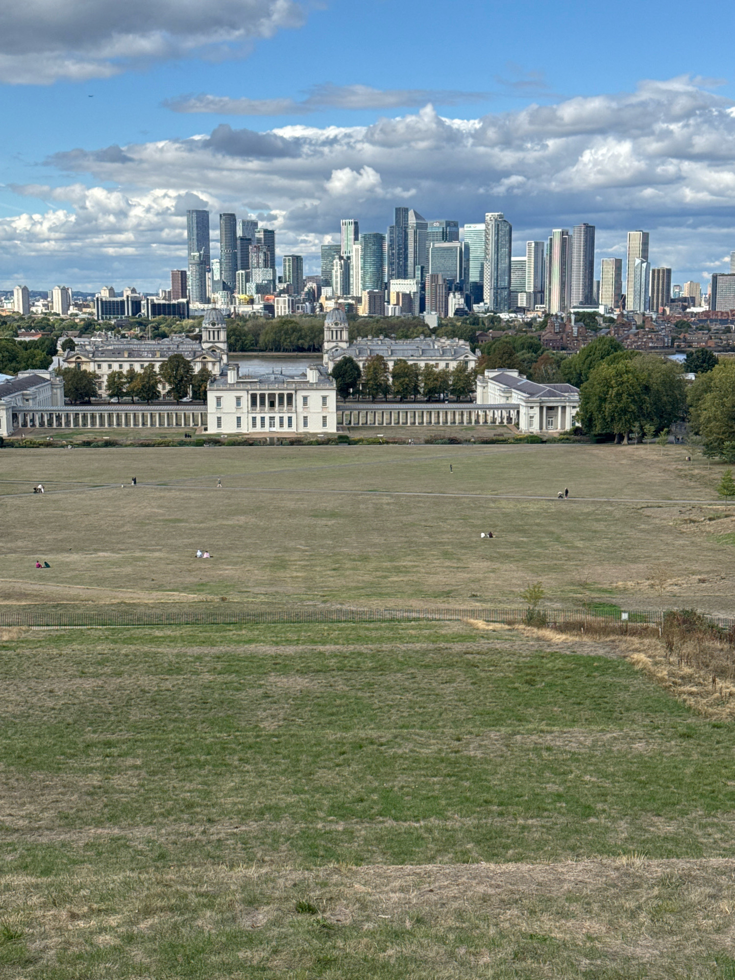 The image depicts a wide-angle view of Greenwich Park in London, United Kingdom. In the foreground, a grassy field with patches of dry, brown grass stretches into the middle ground. Several individuals are scattered across the field, some walking, others sitting or lying down. A wooden fence runs horizontally across the middle ground, separating the field from a paved pathway behind it.
In the middle distance, a large, white, classical-style building with a colonnade is visible. Behind this building, the River Thames can be seen, and beyond that, the modern skyline of Canary Wharf, characterized by numerous tall skyscrapers, dominates the background. The sky is blue with scattered white clouds. The overall scene suggests a pleasant day in a park with a striking juxtaposition of historic architecture and a contemporary urban landscape.