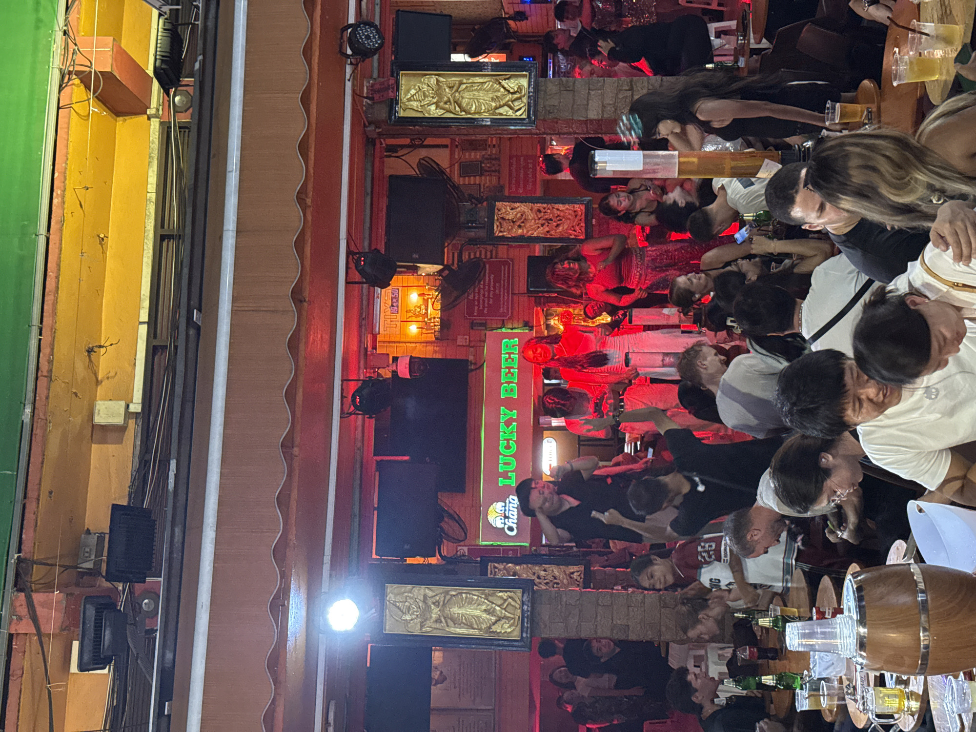 The image depicts a bustling bar scene in Bangkok, Thailand. A crowd of people, predominantly tourists, are densely packed around tables, many holding drinks and engaging in conversations. The bar, named "LUCKY BEER" as indicated by a prominent neon sign, is brightly lit with red lighting. Speakers and golden artifacts are mounted on the walls. The external facade shows a mix of architectural styles with wooden and metal framework. Patrons are leaning close to each other, suggesting a lively and potentially loud environment.