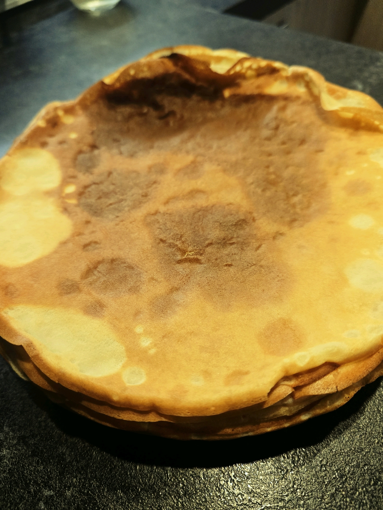 A close-up, overhead shot captures a stack of golden-brown crepes resting on a dark, textured surface. The crepes exhibit varying degrees of browning, with some areas lighter and others darker, indicating they have been recently cooked. The stack is slightly askew, with the top crepe curving upwards. The background is out of focus, showing a dark, possibly kitchen counter, environment. The image does not provide any information about the city of Błonie, Poland.