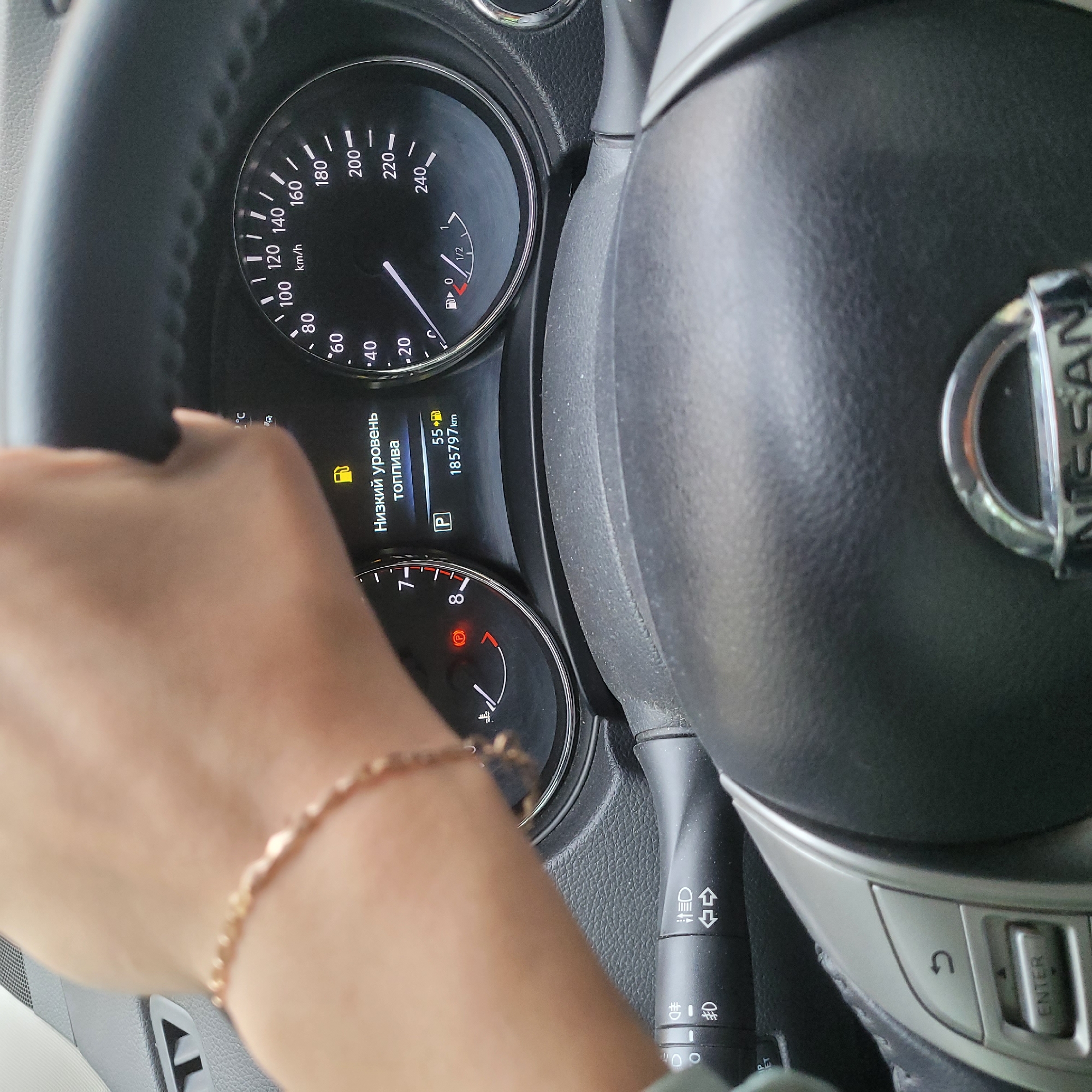 A first-person perspective shot shows a hand with a gold bracelet gripping the steering wheel of a Nissan. The dashboard displays a low fuel warning in Russian, reading "Низкий уровень топлива" (Low fuel level), along with the mileage of 185797 km. The speedometer is visible, with the needle pointing to approximately 60 km/h. Other indicators on the dashboard include a temperature gauge and a parking brake light. The steering wheel has control buttons on the left side, including a "back" button and an "enter" button. The environment appears to be inside a car in an urban setting, though no specific city or location details are discernible.