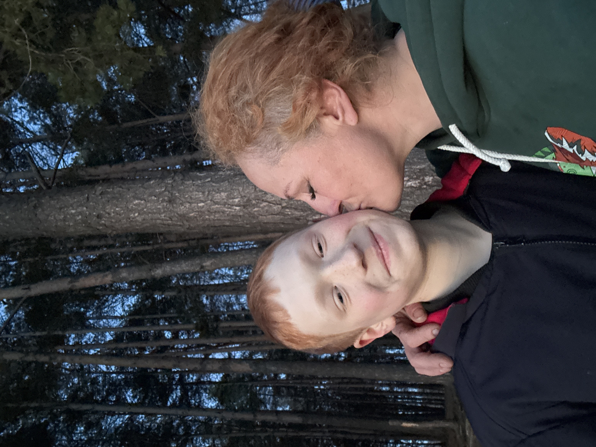 An intimate moment between two individuals is captured in a photo rotated 90 degrees counter-clockwise, set in what appears to be a forest in Chaikovsky, Russia, during twilight hours.
The main subjects are an adult woman and a child, both with reddish-blonde hair. The woman, positioned on the left, has curly hair and is wearing a dark green hoodie with white drawstrings. A colorful graphic resembling an animal or monster face with sharp teeth is partially visible on her hoodie. She is tenderly kissing the child's forehead, her eyes closed in the affectionate gesture.
The child, on the right, has a fair complexion with freckles across their nose and cheeks. They are wearing a dark blue or black zipped jacket over a bright red garment and are looking towards the viewer with a soft, content smile. Their hand is visible near their chest.
The background consists of numerous dark, vertical tree trunks, indicative of a dense wooded area, possibly a pine forest. Through the gaps in the trees, a deep blue sky is visible, suggesting the photograph was taken at dusk or early evening, creating a serene and peaceful atmosphere. There is no visible text in the image.
