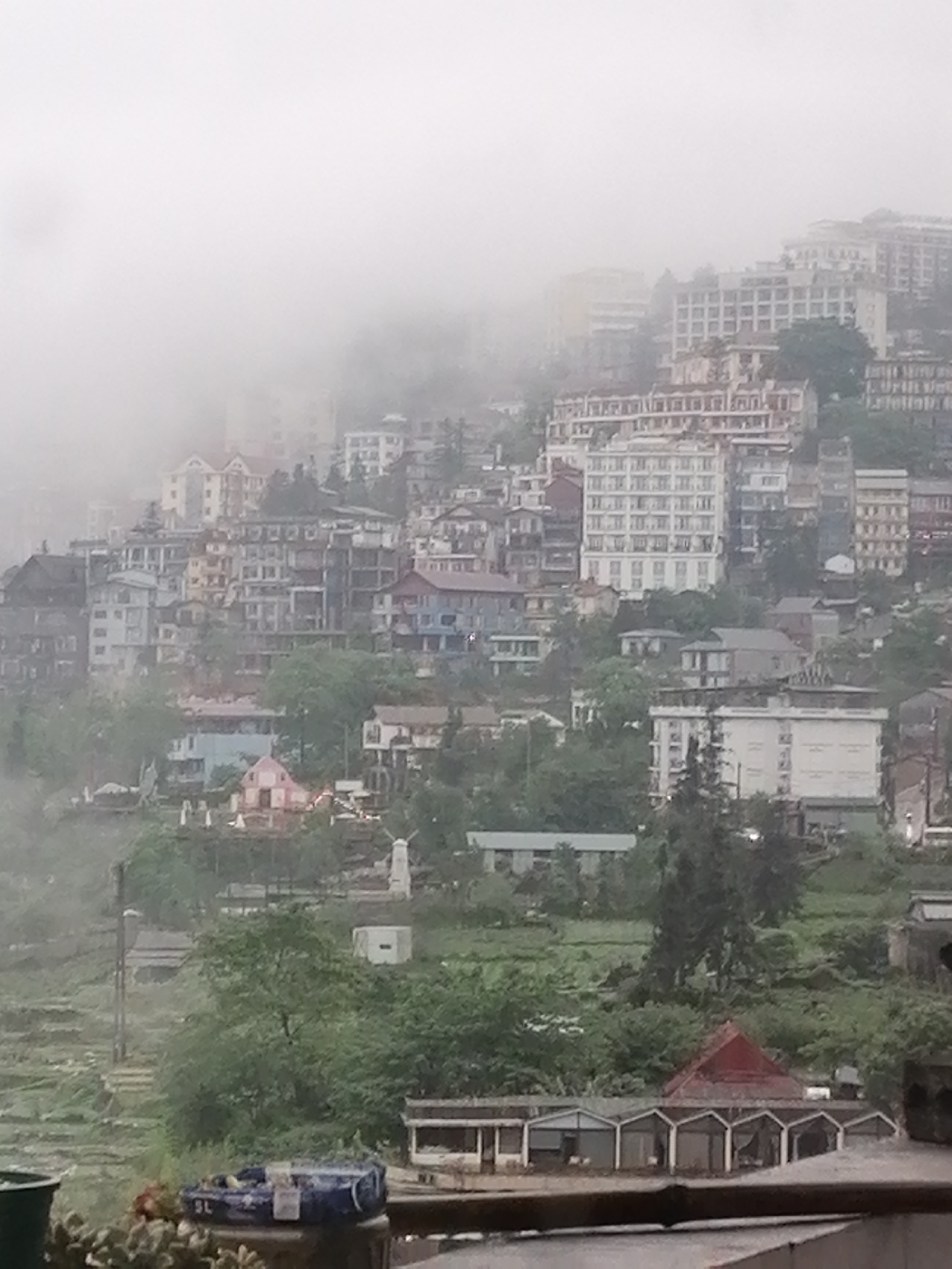 An overview of the built environment within Sa Pa Ward, Vietnam, reveals a multi-tiered urban landscape densely populated with buildings of varying heights and architectural styles. Buildings of up to ten stories are perched on hillsides, many partially obscured by heavy fog. The infrastructure includes residential and commercial structures, which appear to be of modern construction. The foreground features balcony objects, like pots and small shelters. The scene is predominantly gray and green due to the overcast weather and the vegetation interspersed among the buildings. Overall, the scene depicts the densely built environment of Sa Pa Ward under foggy weather.