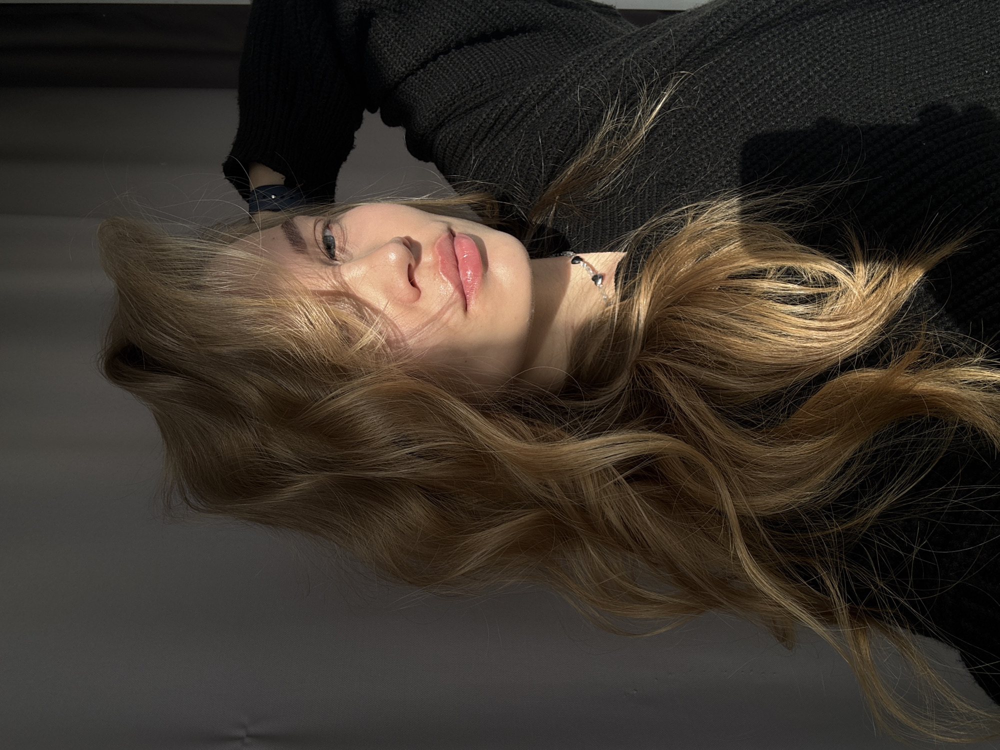 A woman with blonde hair is lying down, wearing a black sweater and a silver necklace. Part of her face is illuminated by sunlight. The background is a grey surface. The location is Chaikovsky, Russia.