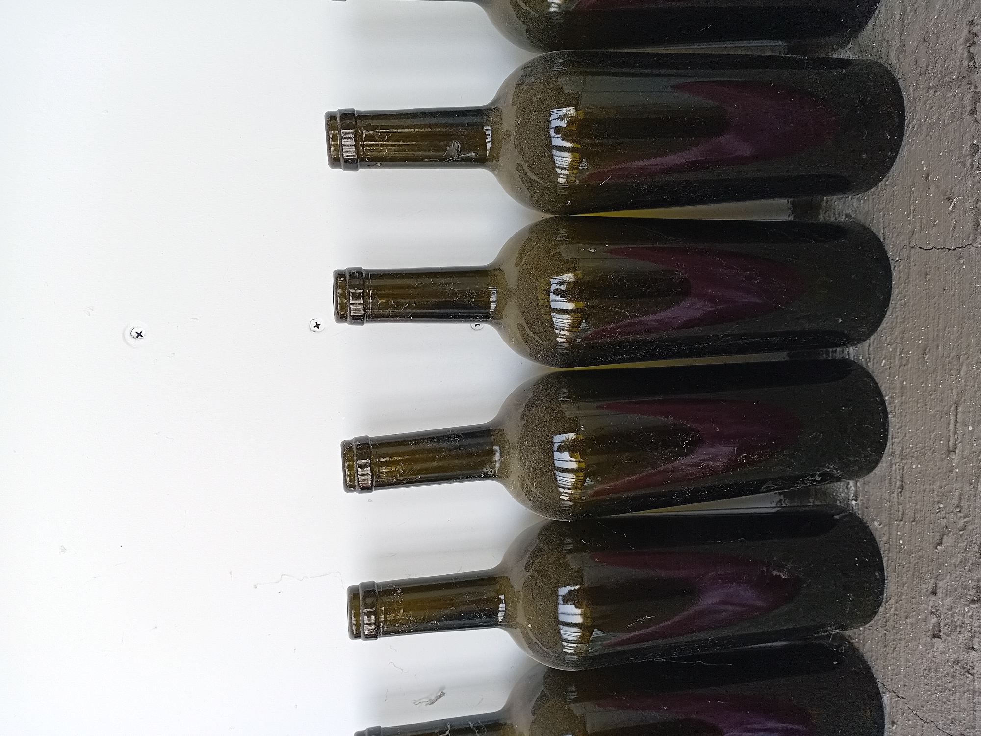 A line of empty, dusty, dark green glass bottles stands upright against a white wall. The bottles are positioned on a rough, textured grey surface, possibly concrete, which is also speckled with dust. The wall behind the bottles has some faint lines and two small screws visible. The bottles have a reflective surface, showing a distorted reflection of what appears to be a person holding a camera and a window or building. The image is taken at an eye-level perspective. The location is identified as Akhasheni, Georgia, a region known for its wine production.