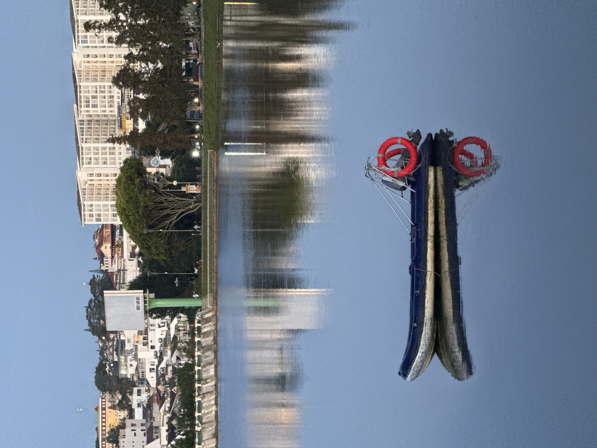 The image depicts Xuan Huong Lake in Da Lat, Vietnam. A dark blue inflatable boat with a white stripe along the bottom is moored in the calm water, its reflection clearly visible. A red life preserver is attached to a metal frame at the stern of the boat. In the background, the city of Da Lat rises up a hill. Buildings of various sizes and architectural styles, including a large modern white apartment complex, are visible. A large billboard stands prominently on the left side of the background. Trees line the shore of the lake and intersperse among the buildings. The sky is a clear, pale blue, indicating either early morning or late evening light. The overall scene is peaceful and serene, capturing a quiet moment on the lake.