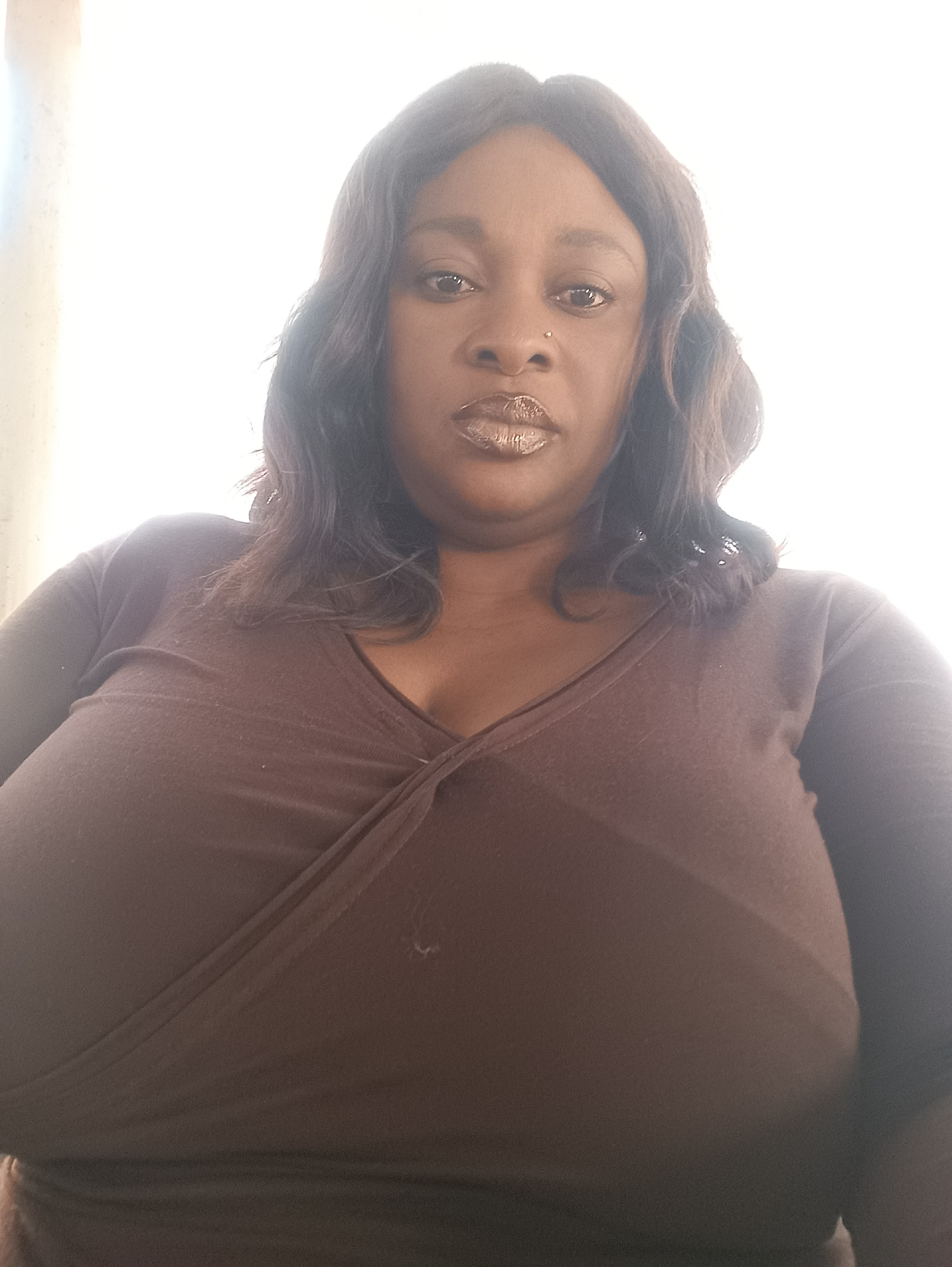 A medium-shot, point-of-view photograph depicts a woman with dark skin and wavy, dark hair. She is wearing a brown v-neck top that appears to wrap or cross in the center of her chest. Her face is centrally framed; she has dark eyes, full lips with a metallic-toned lip color, and a nose piercing in the left nostril. The background is largely out of focus and appears to be a brightly lit, possibly outdoor, setting.
No information within the image indicates a specific location in the city of Jos, Nigeria.