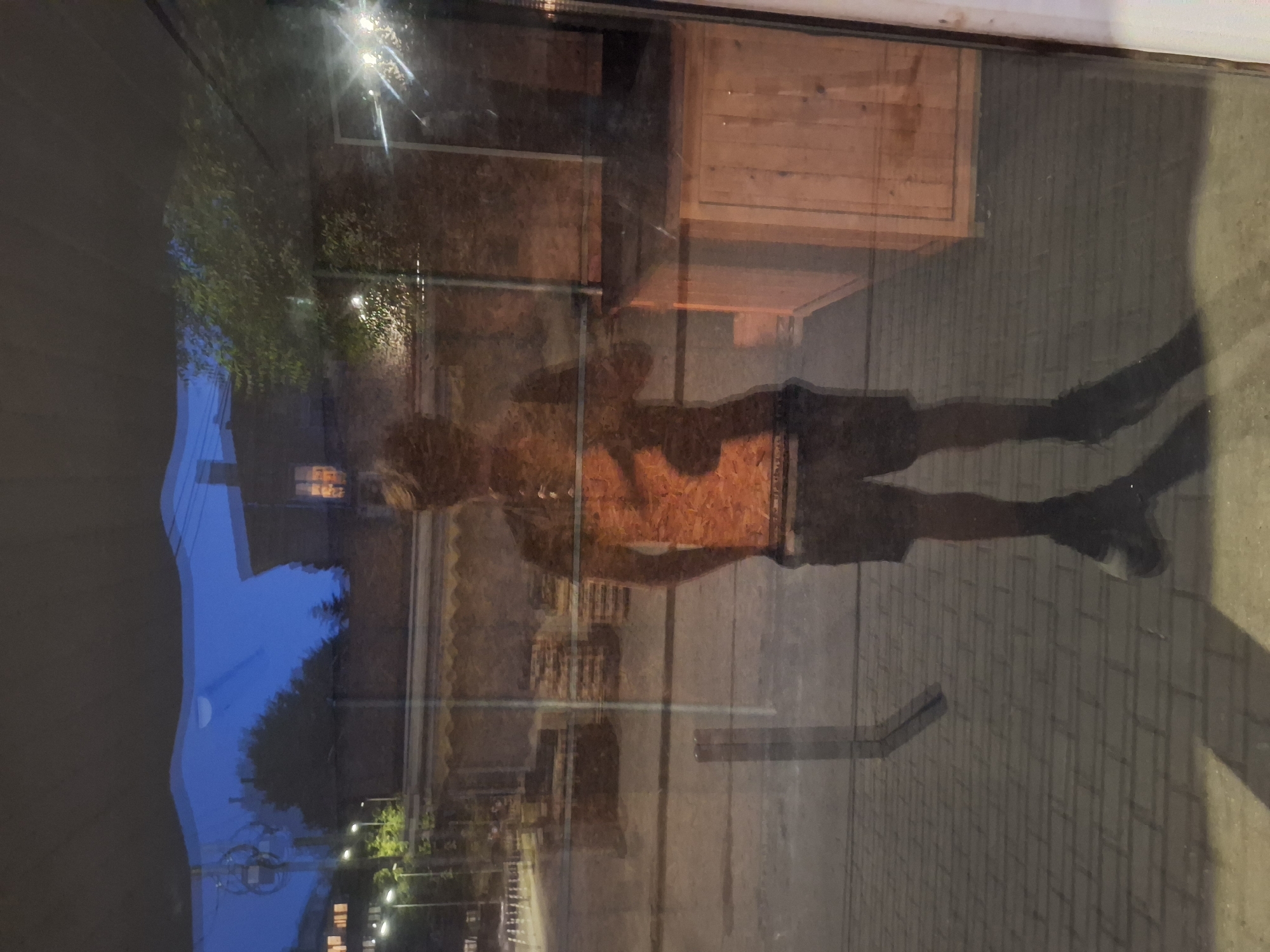 A full-body reflection of a person is visible in a large pane of glass, likely a shop window. The individual is wearing dark shorts, a light-colored tank top with a patterned design, and dark shoes. They are standing on a paved surface. The background, seen through the glass, depicts a street scene at dusk. Buildings with illuminated windows are visible, along with streetlights and trees. The overall atmosphere suggests an urban environment in Varna, Bulgaria. The reflection is clear, with the surroundings subtly distorted by the glass.