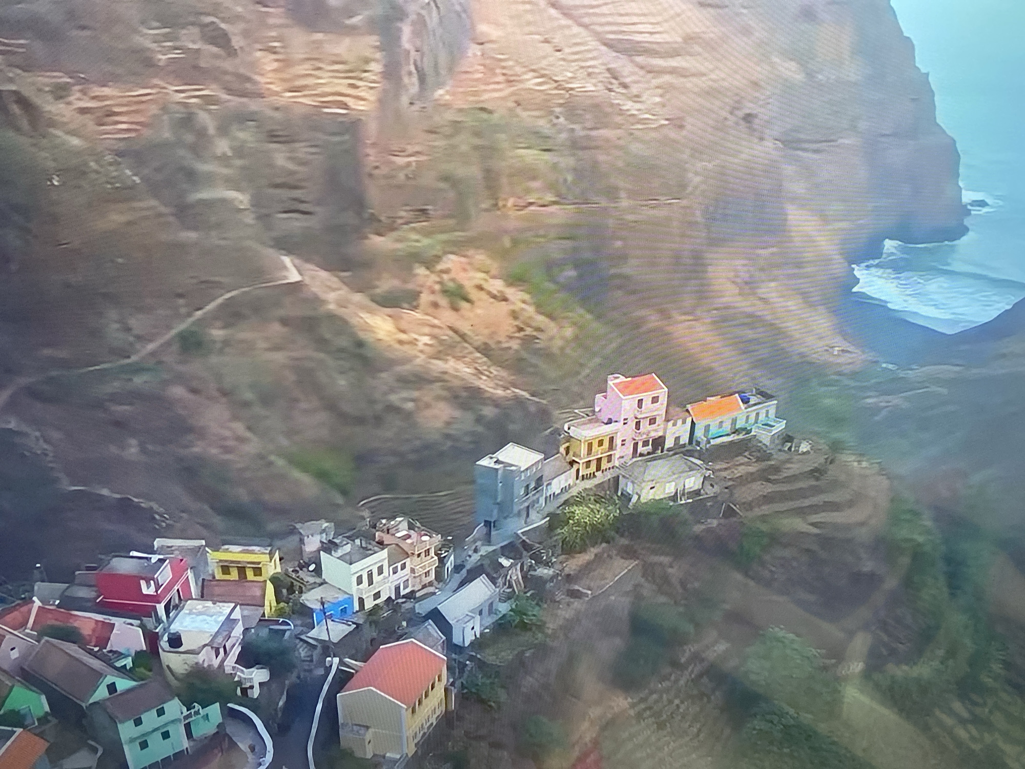An aerial, oblique view captures a colorful village nestled precariously on a steep, terraced mountainside that descends towards a turquoise ocean. The village consists of numerous small, brightly painted houses, predominantly in shades of pink, yellow, blue, and red, with white accents. The structures are tightly packed, following the contours of the terraced landscape, and appear to be constructed from concrete or stucco. Some houses have visible water tanks on their roofs. A narrow, winding path is visible on the mountain above the village. The coastline is characterized by sheer cliffs and rough surf. The overall impression is one of a remote, vibrant settlement existing in a dramatic natural environment. This scene does not depict the city of Minsk, Belarus.