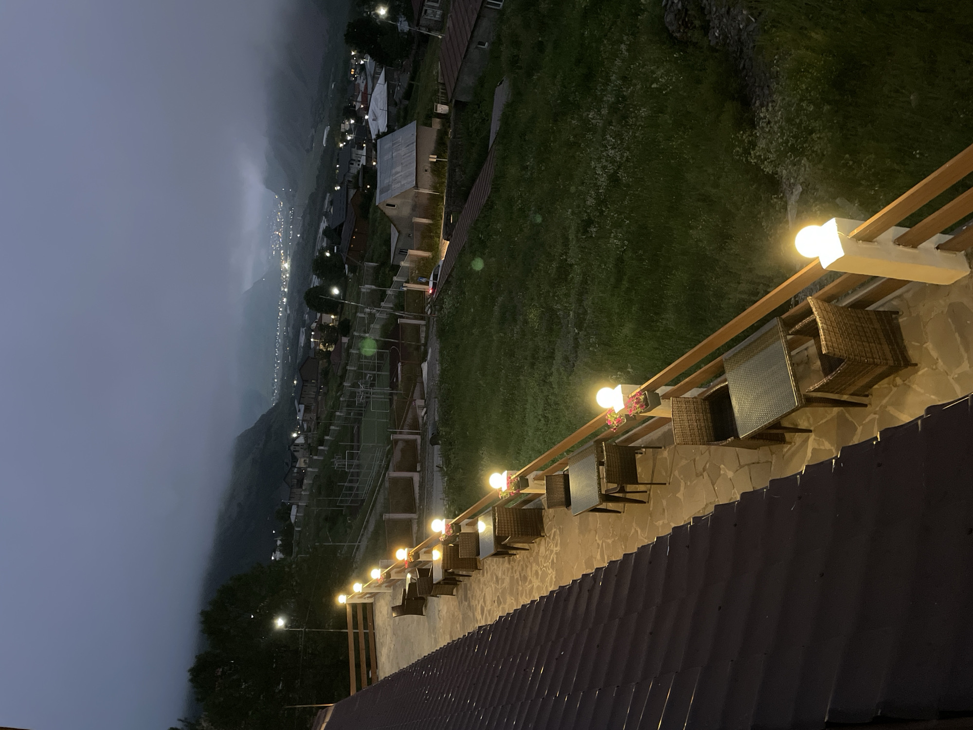 This image depicts an outdoor seating area of a restaurant or cafe in Stepantsminda, Georgia, at dusk or early night. The foreground shows a dark, sloped roof, likely made of dark metal shingles. To the right of the roof, a paved patio area features several outdoor tables and chairs made of wicker-like material. These tables are set with what appear to be glass-topped surfaces. The patio is bordered by a wooden railing, along which are posts topped with illuminated spherical light fixtures. Small flower pots with pink flowers are placed on some of these posts.
Beyond the patio, a grassy slope descends towards a valley. In the mid-ground, several buildings, including houses with pitched roofs, are visible. A sports field or court with fences is also present. In the background, a town or village is nestled in a valley, with numerous lights indicating activity. The scene is set against a backdrop of mountains shrouded in low-hanging clouds, creating a somewhat moody and atmospheric environment. The lighting from the patio lamps illuminates the immediate area, while the distant town lights offer a sense of depth and human presence. No people are visible in the image.