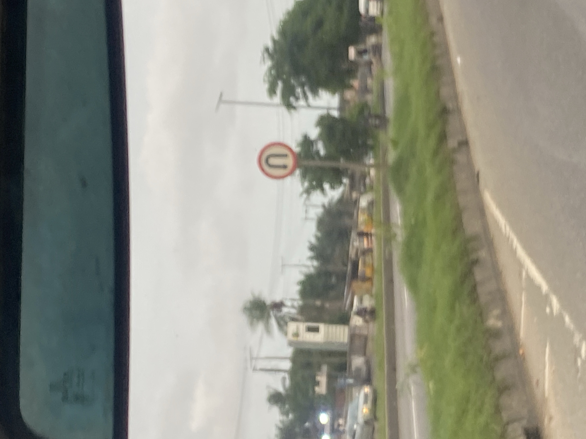The image shows a street scene in Lagos, Nigeria. In the foreground, there is a paved road with a grassy median. In the background, there are several cars, trucks, and motorcycles. There are also trees and buildings. A U-turn sign is visible on the right side of the image. The sky is overcast. The image appears to have been taken from inside a vehicle.