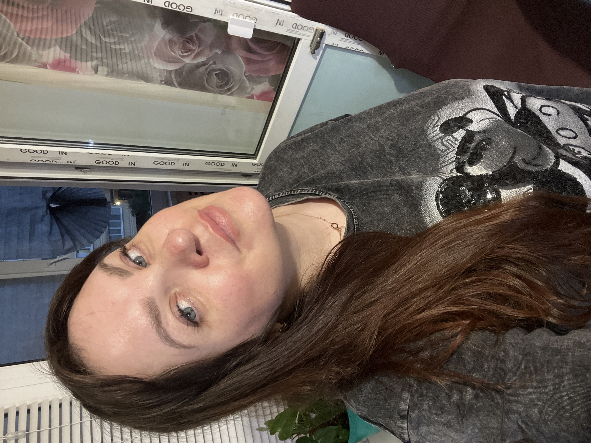 An indoor, selfie-style photograph taken in the city of Noviy, Russia, depicts an adult female with long, brown hair and blue eyes. She presents a faint smile while looking directly at the camera. Her attire includes a grey, short-sleeved t-shirt with a sequined black-and-white graphic design and a gold-toned necklace with small, interlocking circular elements. The background features a white UPVC window frame, vertically marked with "GOOD IN" text. Through the window, on the left, white Venetian blinds partially obscure a green plant in a turquoise pot, while the exterior is dark, indicating night or deep dusk. On the right, a curtain or blind adorned with large pink and grey rose patterns is visible through the window. The room's interior includes a section of a light blue wall and a fragment of dark red upholstery on the far right.