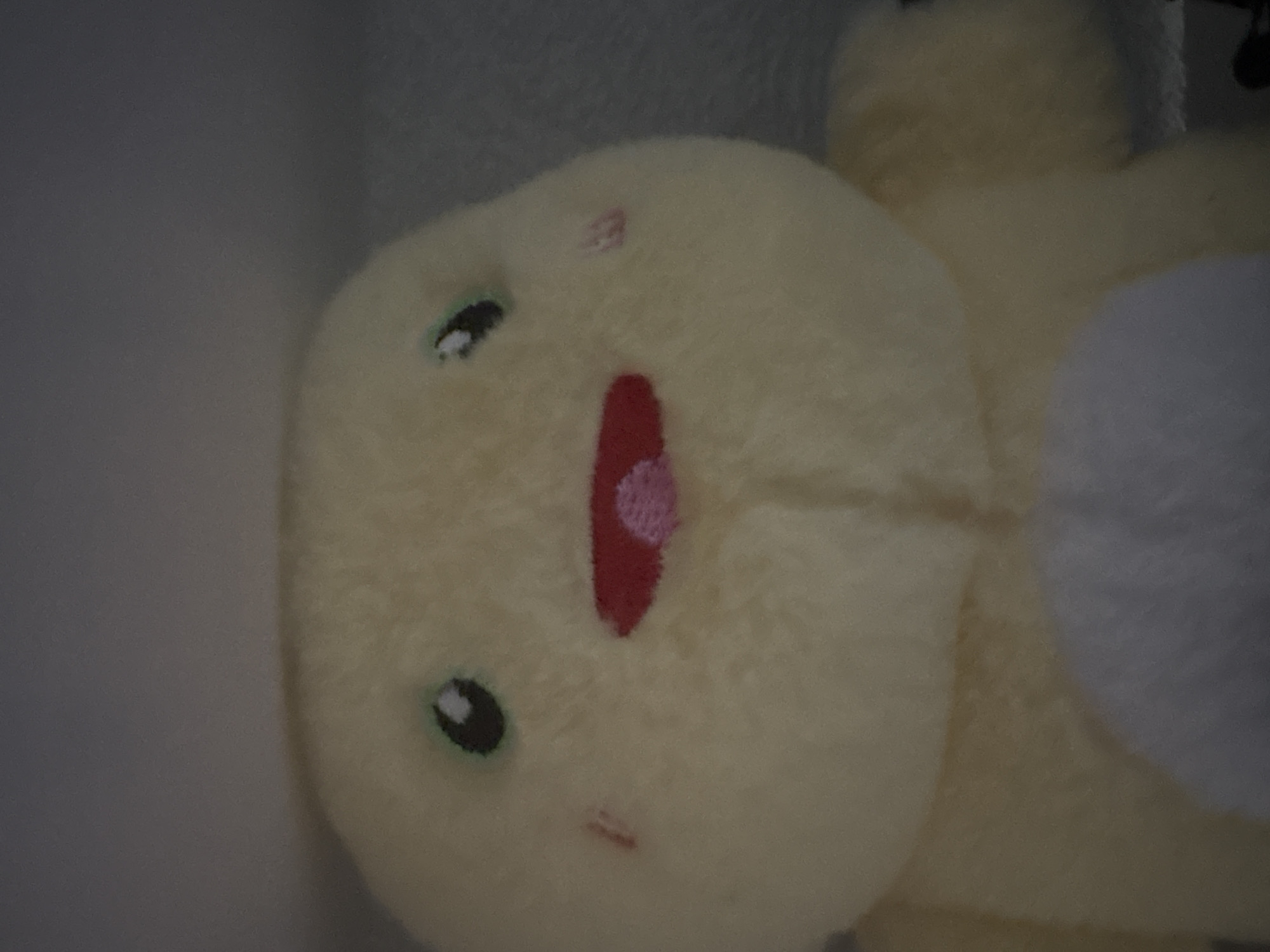 The image is a close-up, slightly blurry photograph of a yellow plush toy, possibly a character from a cartoon or video game. The toy has large, round, dark eyes with green irises and black pupils. It has a wide, red mouth with a pink oval shape inside, suggesting an open mouth with a tongue. Two small pink blush marks are visible on its cheeks. The toy is made of soft, fuzzy material. The background is a muted gray, suggesting an indoor setting, possibly a shelf or a wall. There is no discernible information about the location in the city of Tulinskiy, Russia, as the image does not contain any contextual clues to suggest this geographical placement.