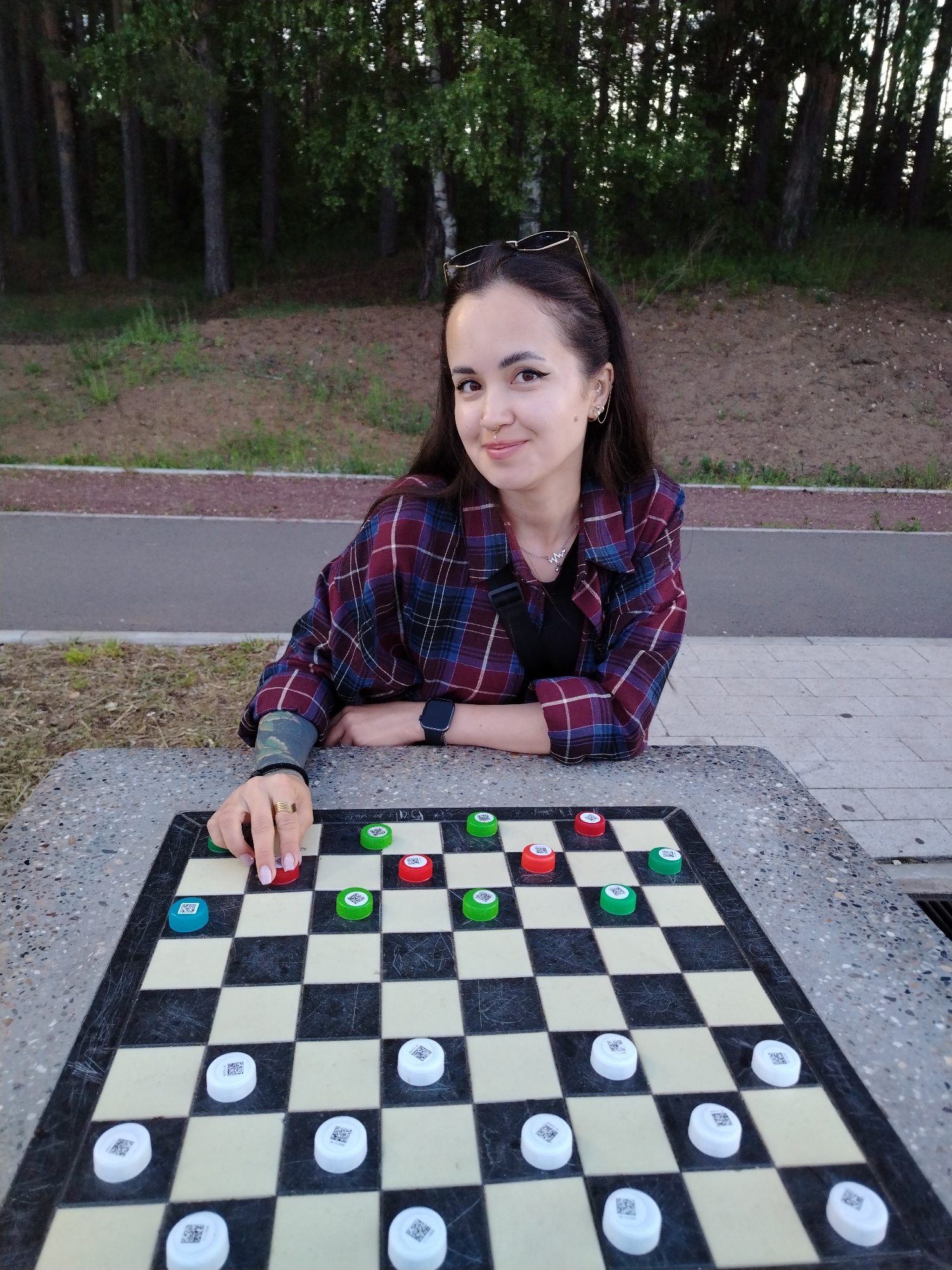 An outdoor scene in Chaikovsky, Russia, depicts a female individual seated at a stone table with an integrated checkerboard. The individual, a young adult with dark hair, a septum piercing, multiple ear piercings, and a mole on her right cheek, wears sunglasses on her head, a plaid shirt (burgundy, dark blue, white, black), a black top, a black shoulder bag strap, a dark smartwatch on her left wrist, and a gold-colored ring on her right ring finger. A camouflage pattern is visible on her right forearm. She is looking directly at the viewer with a slight smile, with her right hand positioned above a red game piece on the board, indicating an active game.
The checkerboard features standard black and off-white squares. Game pieces are unconventional, appearing to be bottle caps: five white pieces with visible QR codes are positioned in the foreground, while seven pieces (a mix of red, green, and one light blue) are positioned in the upper portion of the board.
In the background, a paved asphalt path with a reddish-brown strip runs horizontally, separating the seating area from a grassy embankment. Beyond the embankment, a dense line of trees, including conifers and deciduous trees, forms the distant background, illuminated by natural light suggesting late afternoon or early evening.