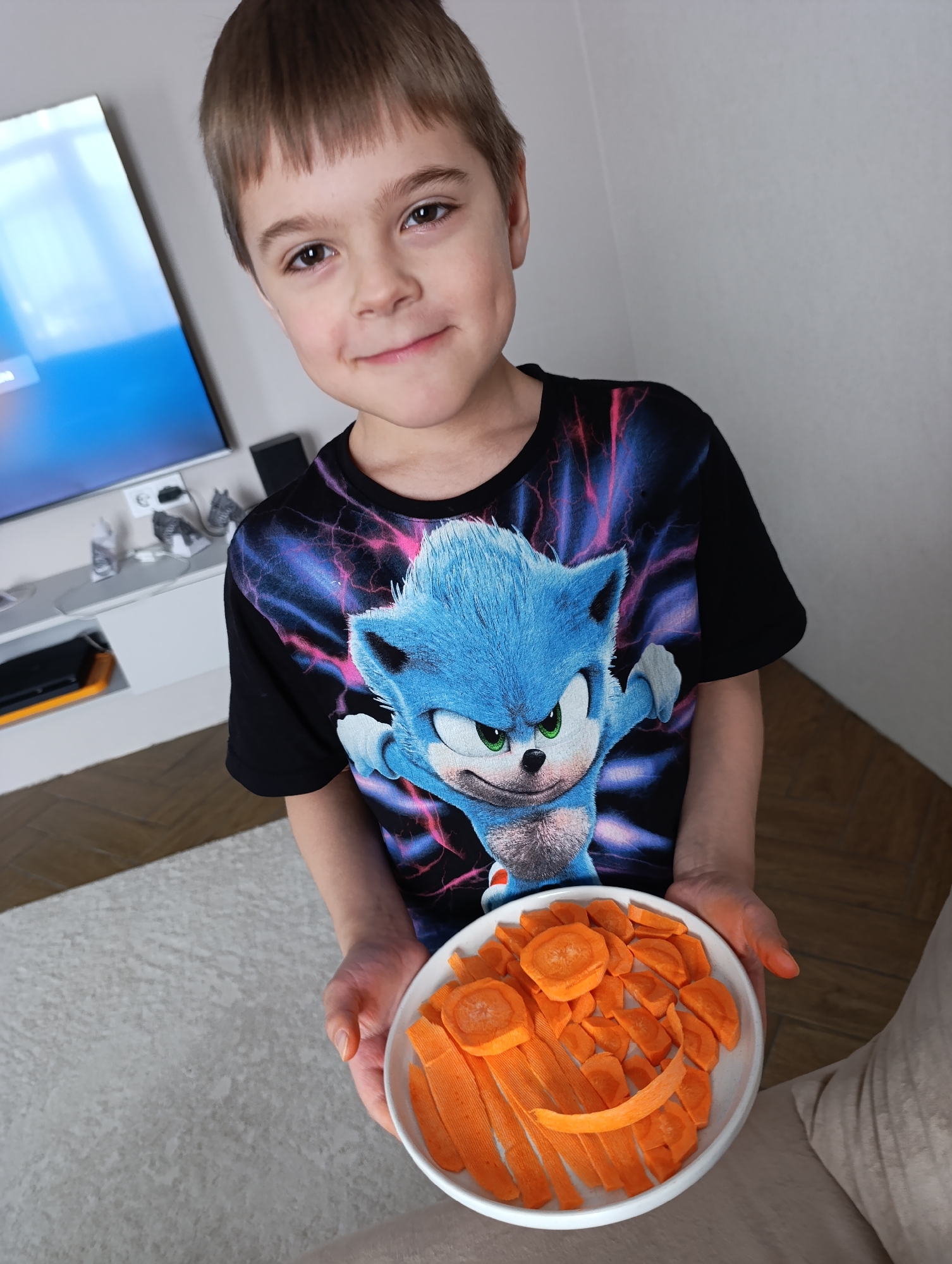 A young boy stands indoors holding a white plate of sliced carrots. He is wearing a black t-shirt with a blue Sonic the Hedgehog graphic on it. The boy has light brown hair and a slight smile. A television is visible in the background along with a console and various other objects. The photo was taken in the city of Chaikovsky, Russia.