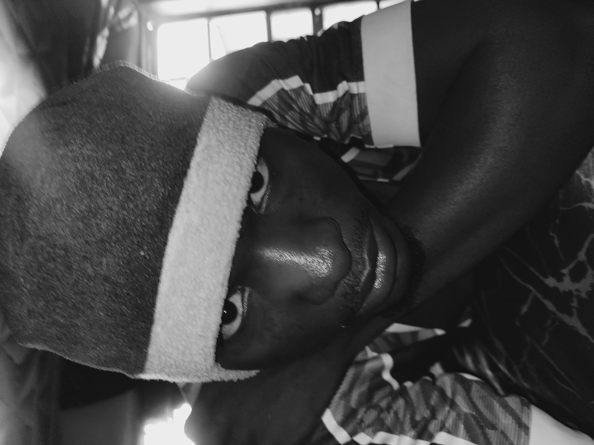 This is a black and white, close-up, low-angle selfie of a young Black male wearing a beanie and a striped shirt. The subject is looking directly at the camera, with his arms crossed and his head resting on his arms. His face is illuminated by a light source from the right, casting shadows and highlighting the texture of his skin. He has a mustache and his lips are slightly parted. The background appears to be indoors, with a window or light source visible to the right. The image was taken in Bukuru, Nigeria.
