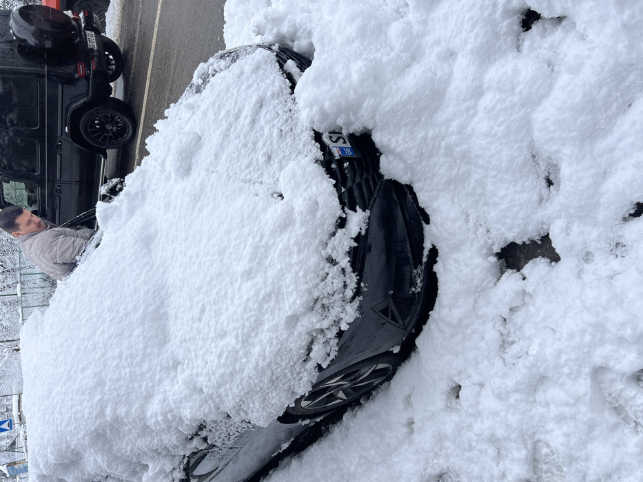 A black car, heavily blanketed in fresh snow, is partially visible. The grill is just exposed, revealing a Georgian license plate. A man sits in the driver's seat of a black SUV parked next to it. The ground and surrounding area are also covered in a thick layer of snow. The image suggests a heavy snowfall event.