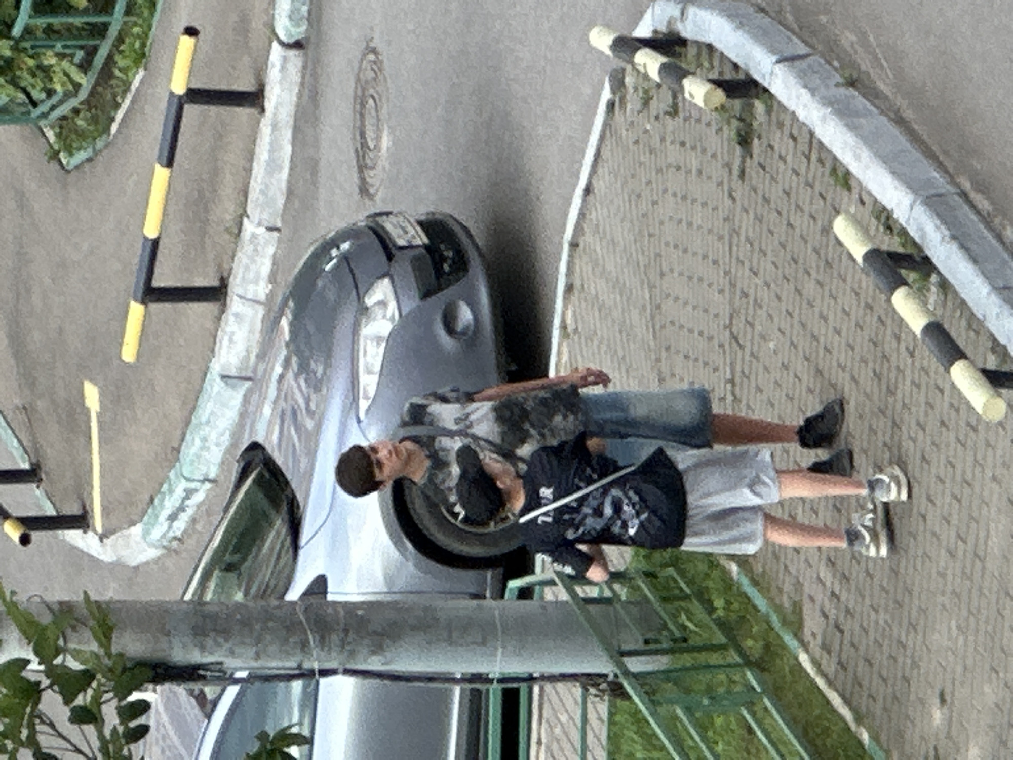 The image depicts two young males standing on a paved walkway adjacent to a street. The male on the left is shorter, wearing a black t-shirt with a graphic design, grey shorts, a black baseball cap, and white sneakers. He has a white crossbody bag over his right shoulder. The taller male on the right is wearing a t-shirt with a camouflage pattern, denim shorts, and dark shoes. He is carrying a dark bag over his left shoulder.
Behind the two males is a gray car parked on the street. The walkway is bordered by a low green metal fence and a concrete curb. The street has a manhole cover visible. In the background, there are black and yellow striped bollards and trees. The overall environment suggests an outdoor urban setting, possibly a residential area in Podolsk, Russia, given the context. The lighting indicates it is daytime.