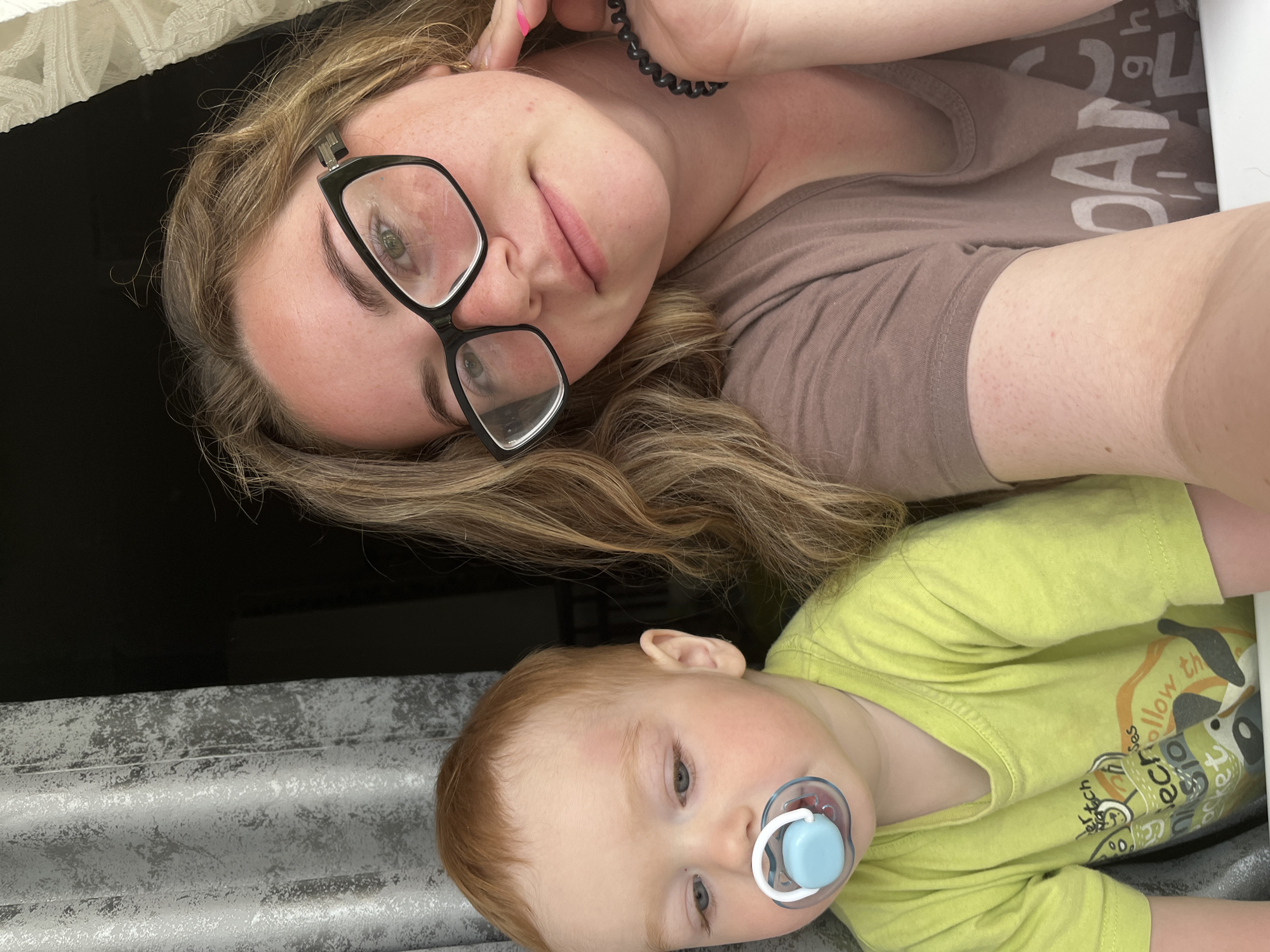 A woman and a baby are positioned together in the image. The woman is wearing glasses and a brown t-shirt and has blonde hair. She is smiling. The baby has a pacifier in his mouth and a yellow t-shirt. They are near a window, with a dark background visible through it. The location is Chaikovsky, Russia.