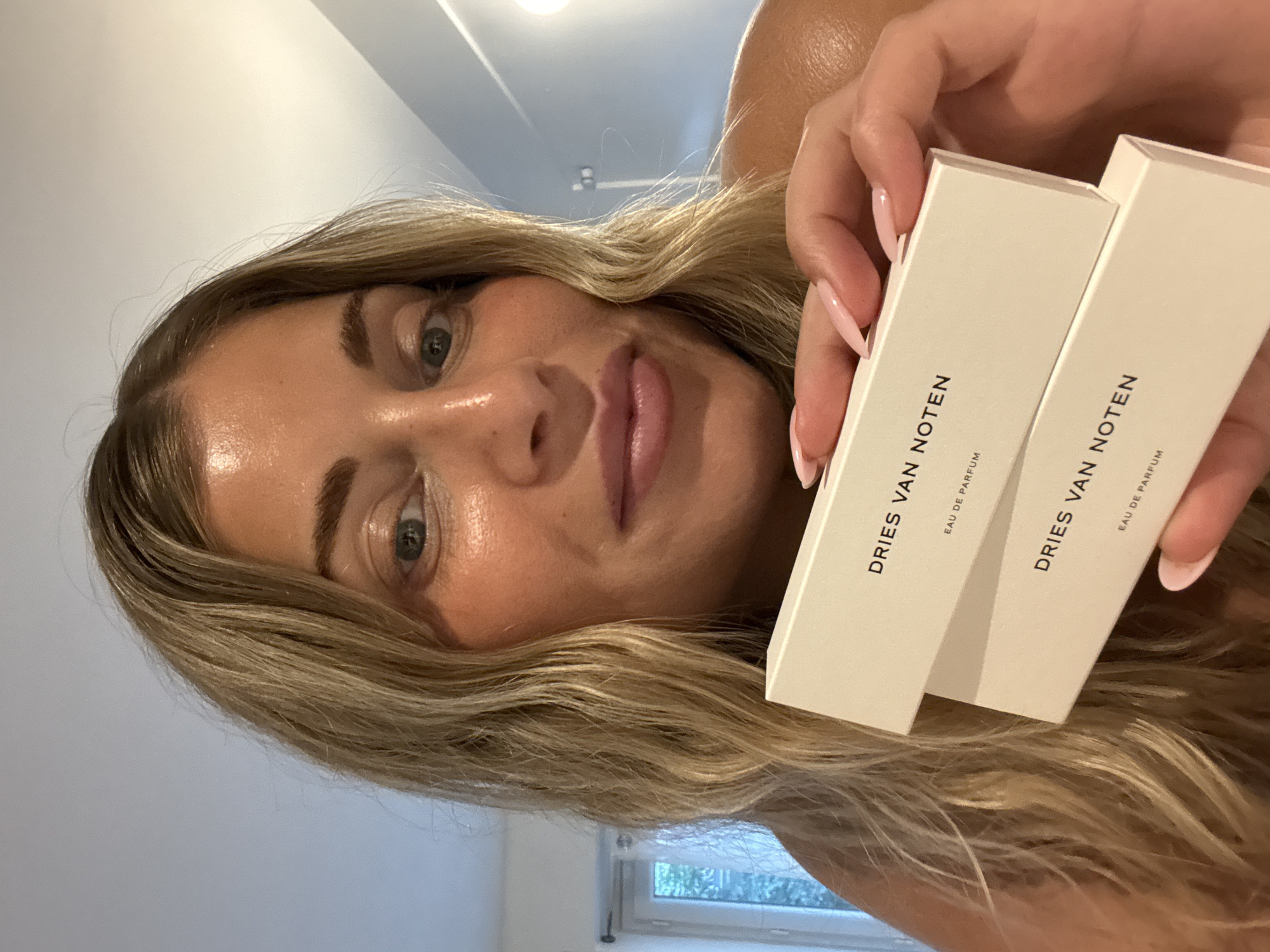 A blonde woman, with her face close to the camera, is holding two identical white boxes of Dries Van Noten Eau de Parfum. The background shows a room with a window. The location in the city of Paris, France is not evident from the image.
