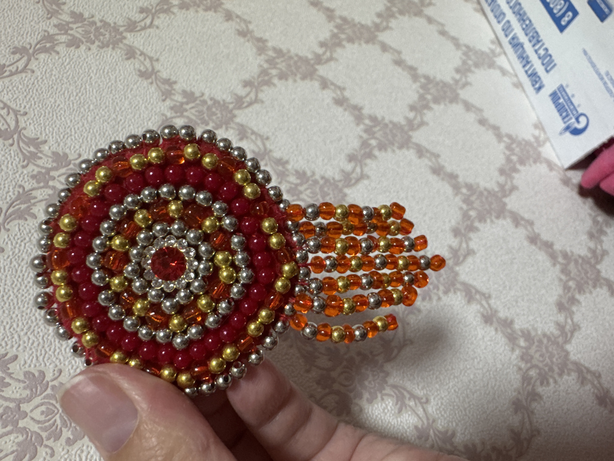 A close-up, vertical shot captures a handmade beaded ornament in the process of being held by a person's thumb and index finger. The ornament is circular, featuring concentric rings of red, gold, and silver beads. A central red gem is surrounded by smaller clear rhinestones, with further rings of beads radiating outwards. Dangling from the bottom edge of the ornament are several strands of smaller orange and gold beads. The background is a textured wallpaper with a subtle damask pattern, rendered in muted tones of beige and grey, with shadows cast on it. In the lower right corner of the image, a partially visible white box with blue text is discernible, featuring the Gazprom logo and the Russian text "КВИТАНЦИЯ ПО ОПЛАТЕ ПОСТАВЛЕННОГО" and "8 (800)". The image does not provide any information about the specific location of Urazovo, Russia, as it is a close-up of an object and its immediate surroundings.