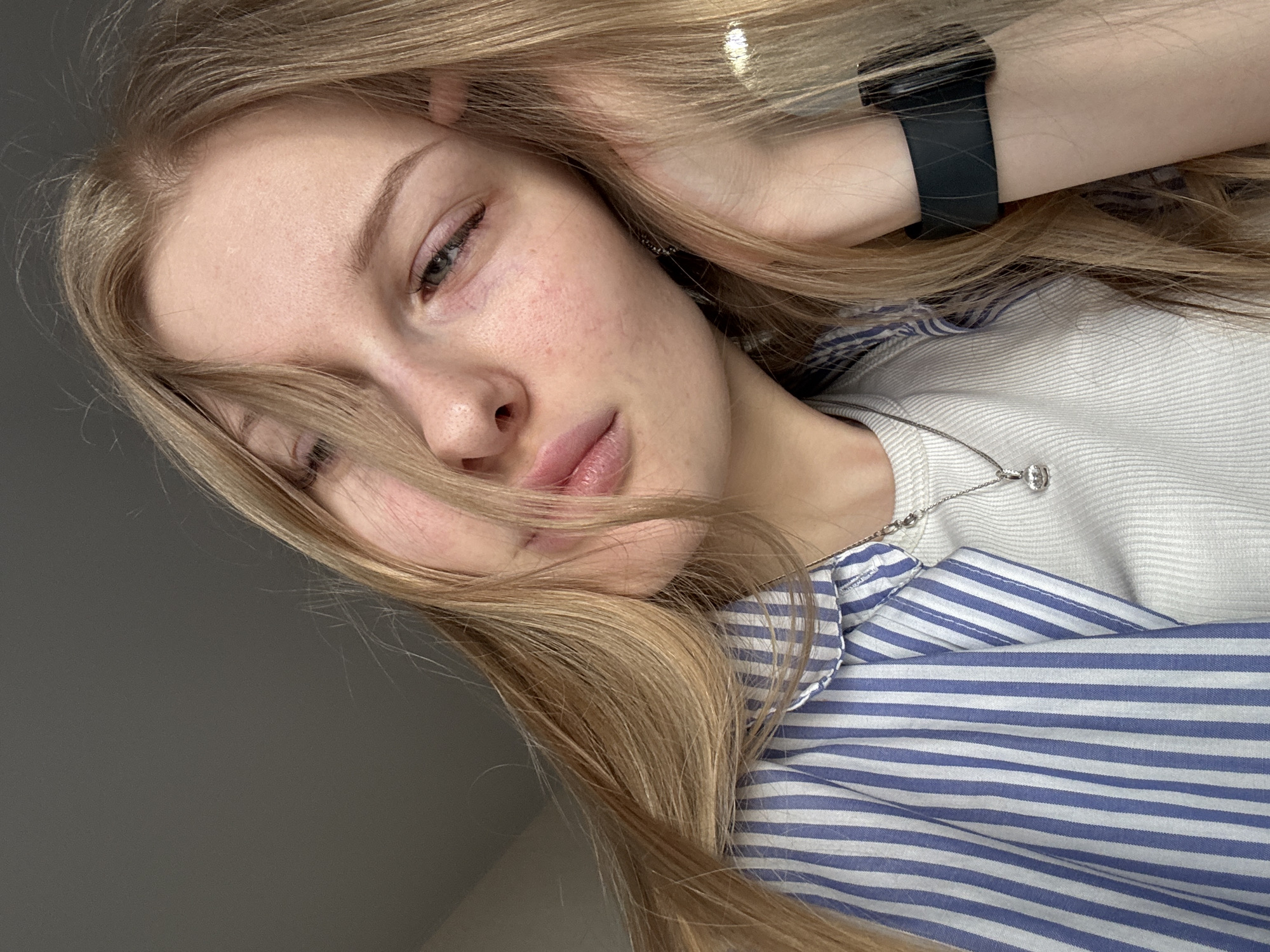 A selfie-style portrait of a fair-skinned young woman with long blonde hair, posed at an angle. She is wearing a white, ribbed top layered under a blue and white striped collared shirt. A black smartwatch is visible on her left wrist. A silver necklace with a pendant is around her neck. Her left hand is raised, with her fingers touching her hair. The background appears to be a plain grey wall. There is no evidence to suggest the location is in Perm, Russia.