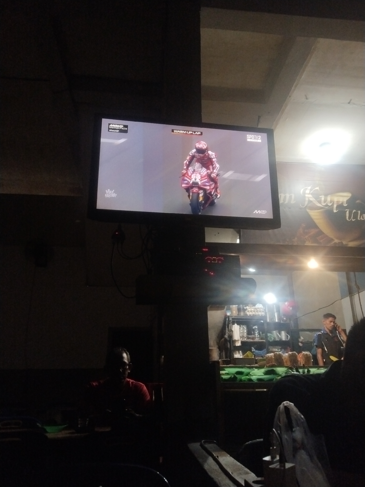 The image depicts an indoor scene, possibly in a public space such as a cafe or eatery. A television screen is mounted high on a wall, displaying a motorcycle race. The broadcast shows a rider on a motorcycle during the warm-up lap.
Below the television, there is a counter area with a person standing behind it, possibly a worker. The person is talking on a mobile phone. There are items on the counter and shelves behind the counter. The lighting in this area is brighter than in the rest of the room.
In the foreground and to the left, there are people sitting at tables, with one person wearing a red shirt visible in the dim lighting. The overall atmosphere suggests a casual setting where people are watching television while possibly eating or drinking.
The location is indicated to be Lhokseumawe, Indonesia.