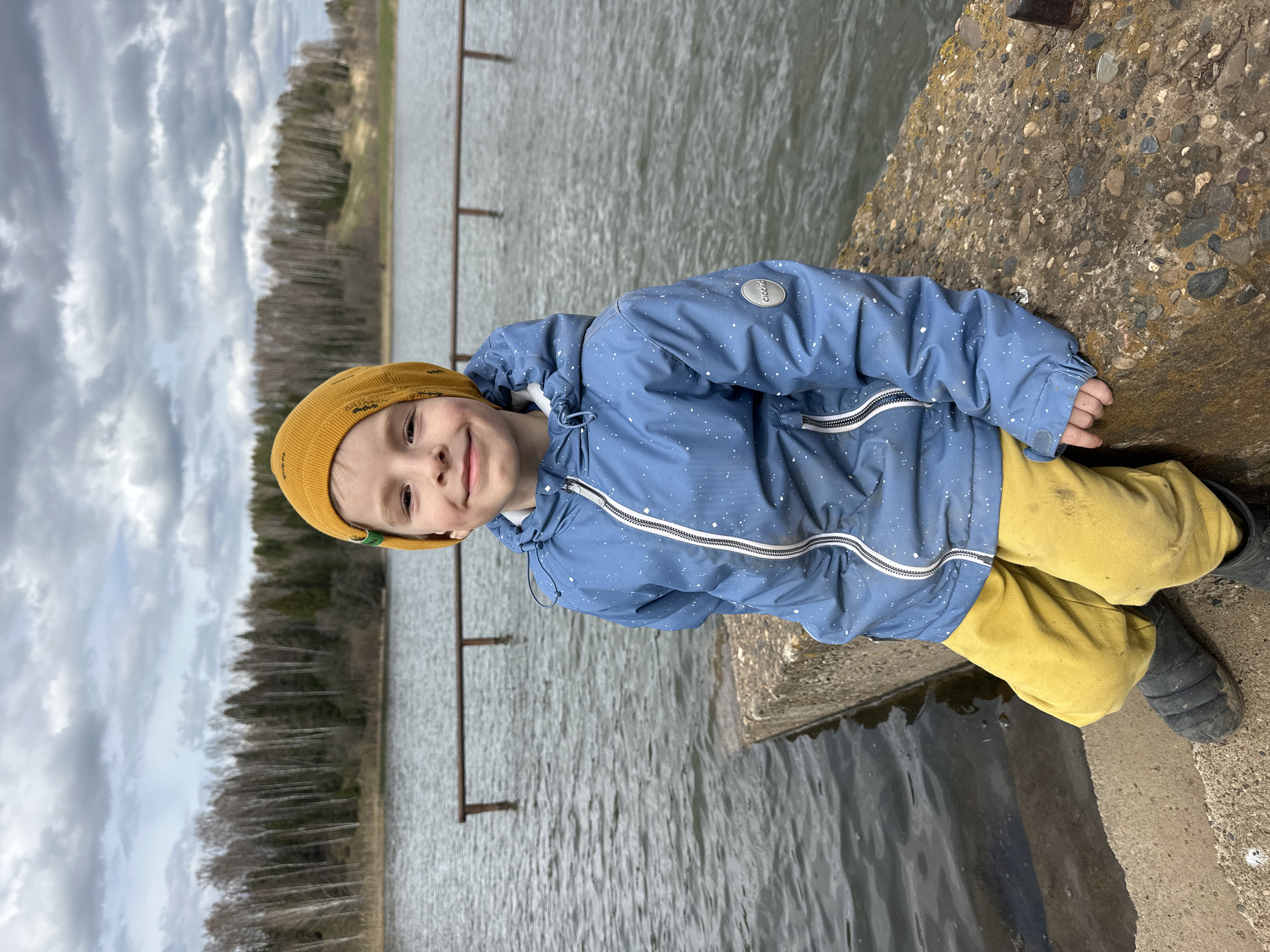 A young boy is smiling and lying on a concrete ledge near a body of water. He is wearing a yellow beanie, a blue jacket with white specks, and yellow pants. The water appears to be part of a lake or reservoir, with metal posts extending from the water's surface. Trees and a cloudy sky are visible in the background. The location is Альняш, Russia.