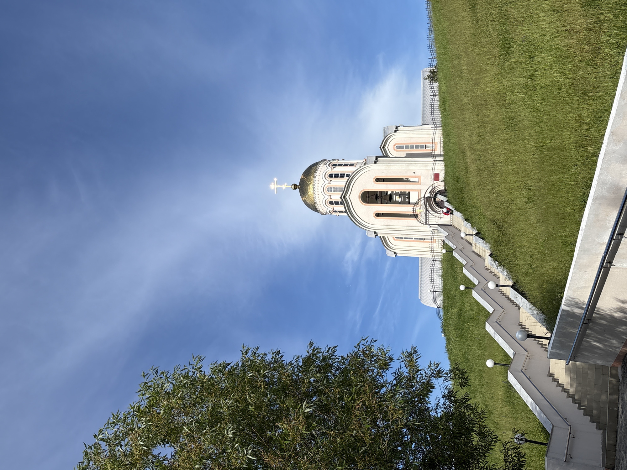 A majestic Eastern Orthodox church stands under a brilliant blue sky, prominently featuring a large, gleaming golden dome topped with a golden Orthodox cross. The church, with its light-colored facade, arched windows, and intricate architectural details, is situated on a verdant hillside covered in lush green grass.
A long, zigzagging concrete staircase with white steps and gray railings ascends the slope towards the church. Spherical light fixtures are regularly spaced along the staircase, suggesting its use after dark. A dark metal fence runs along the top of the grassy hill near the church building. In the lower-left foreground, a tree with dense green foliage partially frames the scene.
The sky is bright and clear with only a few wispy white clouds, indicating a sunny day, likely morning or afternoon, given the strong illumination on the church. One person wearing a red top is visible near the base of the church, adjacent to the staircase. The architectural style and prominent cross are characteristic of Eastern Orthodox churches, aligning with the location context of Varvarovka, Russia. The scene conveys a sense of grandeur and peacefulness. No visible text is present in the image.