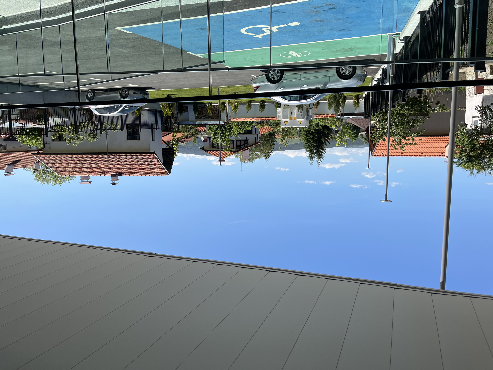 The image depicts a view from an elevated position, showing a reflection on a building's windows of the surrounding area in Belchinski Bani, Bulgaria. In the reflection, one can see residential buildings with red-tiled roofs, green vegetation (trees and shrubs), and a blue sky with light clouds. A parking lot, including a designated handicapped parking space, is also visible in the reflection. The image's foreground shows a gray building facade with vertical lines. A lamppost is visible against the sky. The composition suggests the photograph was taken from inside or near a building with reflective windows, capturing both the exterior view and its reflection.