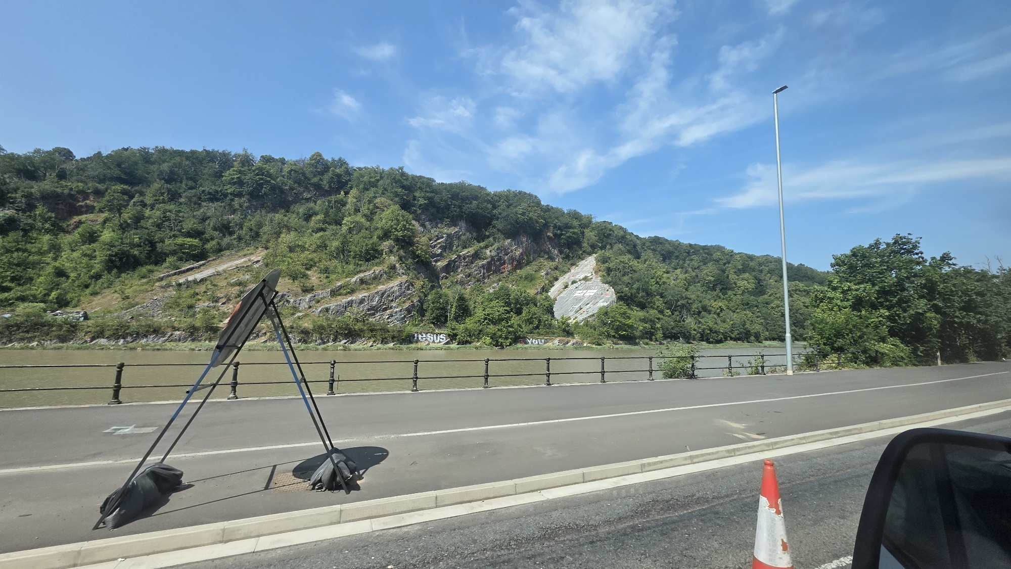 The image depicts a scenic view of the Avon Gorge in Bristol, United Kingdom, looking towards the Clifton Suspension Bridge. The foreground shows a road with a traffic cone and what appears to be a portable barrier or sign. Beyond the road, a metal fence separates the pathway from a body of water, likely the River Avon. On the far bank, a steep, tree-covered hillside is visible, with exposed rock faces. Graffiti is present on the hillside, with the words "JESUS" and "YOU" painted in large white letters. The sky is blue with scattered white clouds. The overall impression is one of a sunny day in a natural setting within the city.