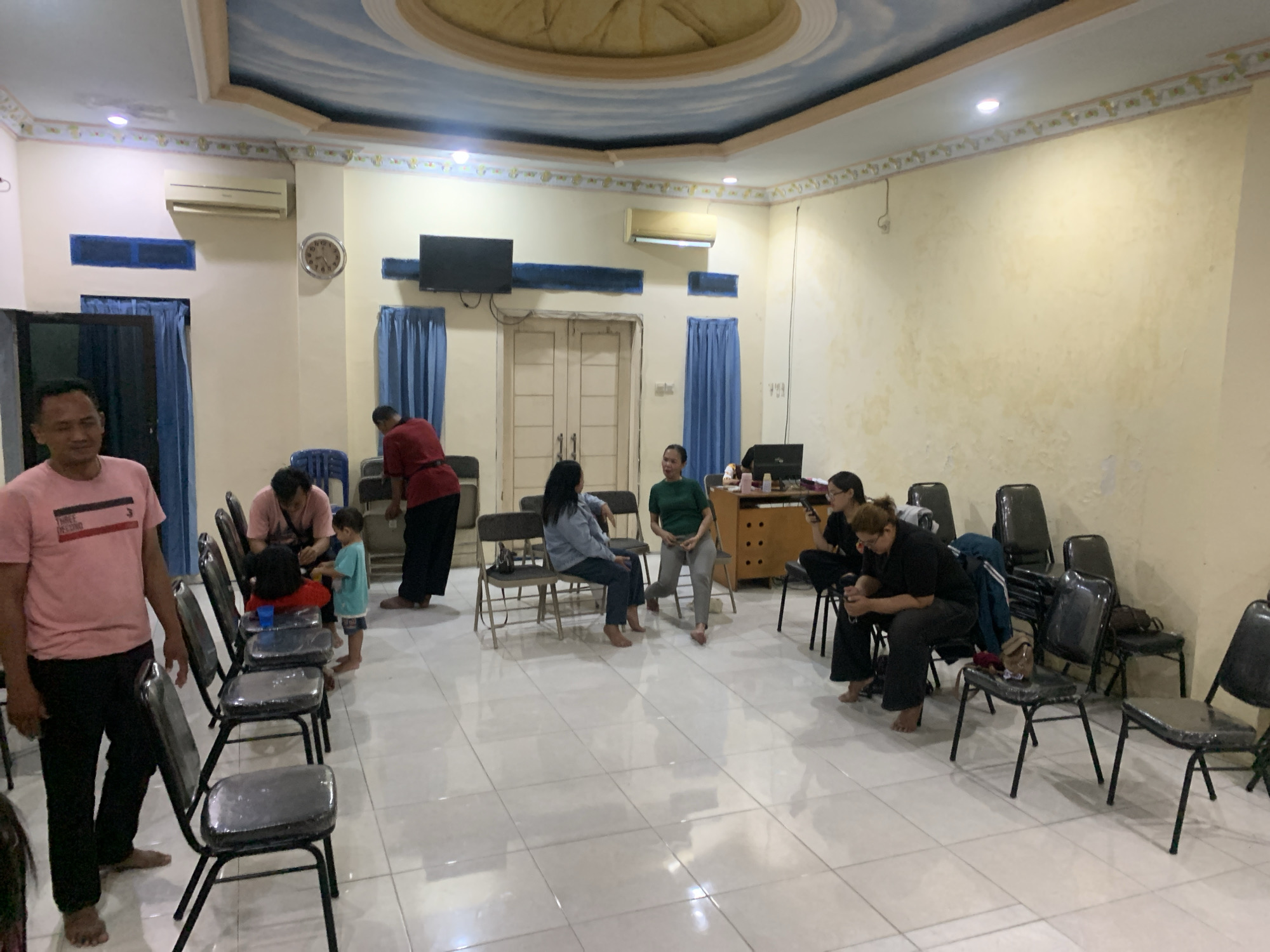 The image depicts an indoor gathering in what appears to be a community hall or meeting room in Dukuh Mojo, Indonesia. Several individuals are seated on chairs arranged in rows, facing the front of the room, although their attention is directed in various directions. The room features a tiled floor, cream-colored walls, and a decorative ceiling with a painted sky mural. Blue curtains hang over windows or doorways, and an air conditioning unit is mounted on the wall. A clock with a visible face is also present on the wall.
In the foreground, a man in a pink t-shirt and black pants stands facing towards the left of the frame, his hands by his sides. To his right, a line of black chairs, some covered in plastic, are occupied by a few people. A man in a light pink shirt and a young child in a blue shirt are near these chairs. Further back, a group of women are seated on chairs, engaged in conversation or looking at their phones. One woman in a green shirt is standing. To the right, a desk or cabinet holds various items, including a computer monitor. Another group of people are seated in chairs on the right side of the room, with some appearing to be looking at their phones. The overall atmosphere suggests a casual meeting or social gathering.
