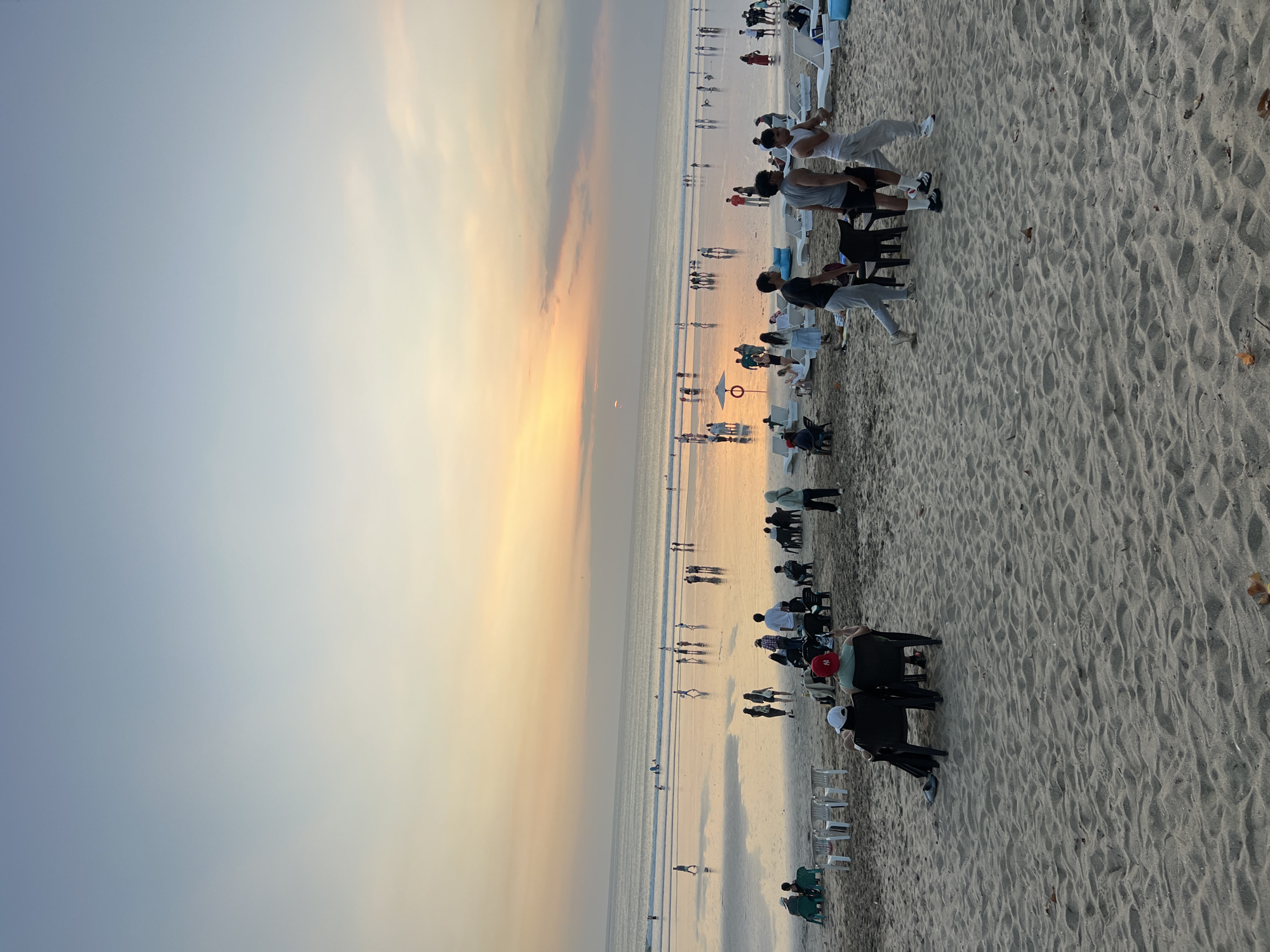 The image depicts a wide-angle view of Kuta Beach, Indonesia, during sunset. The sky is a gradient of pale blue and soft orange hues, with a visible sun low on the horizon. The wet sand of the beach reflects the sky and the distant lights. Numerous people are scattered across the beach and along the water's edge. Some individuals are walking, while others are seated on chairs or lounging on beach chairs in the foreground and mid-ground. The ocean is calm with gentle waves rolling onto the shore. Several small groups of people are visible further out in the water, possibly swimming or surfing. In the foreground, beach chairs and umbrellas are set up. The overall scene is one of a busy but relaxed beach atmosphere at dusk.